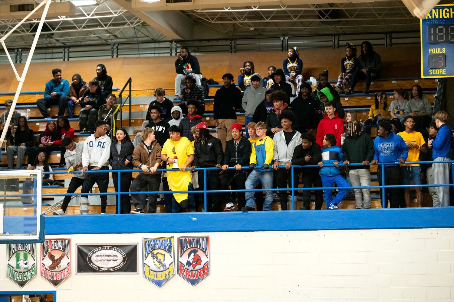Diverse crowd of spectators watches from bleachers at a high school basketball game in a gymnasium.