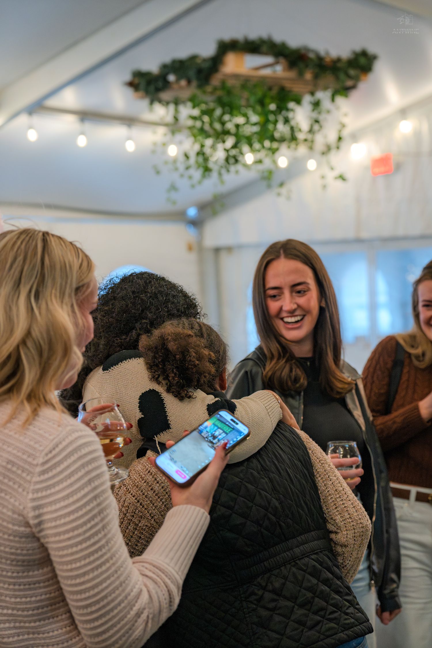 Women laugh and hug at a festive indoor gathering with string lights and greenery hanging from the ceiling.