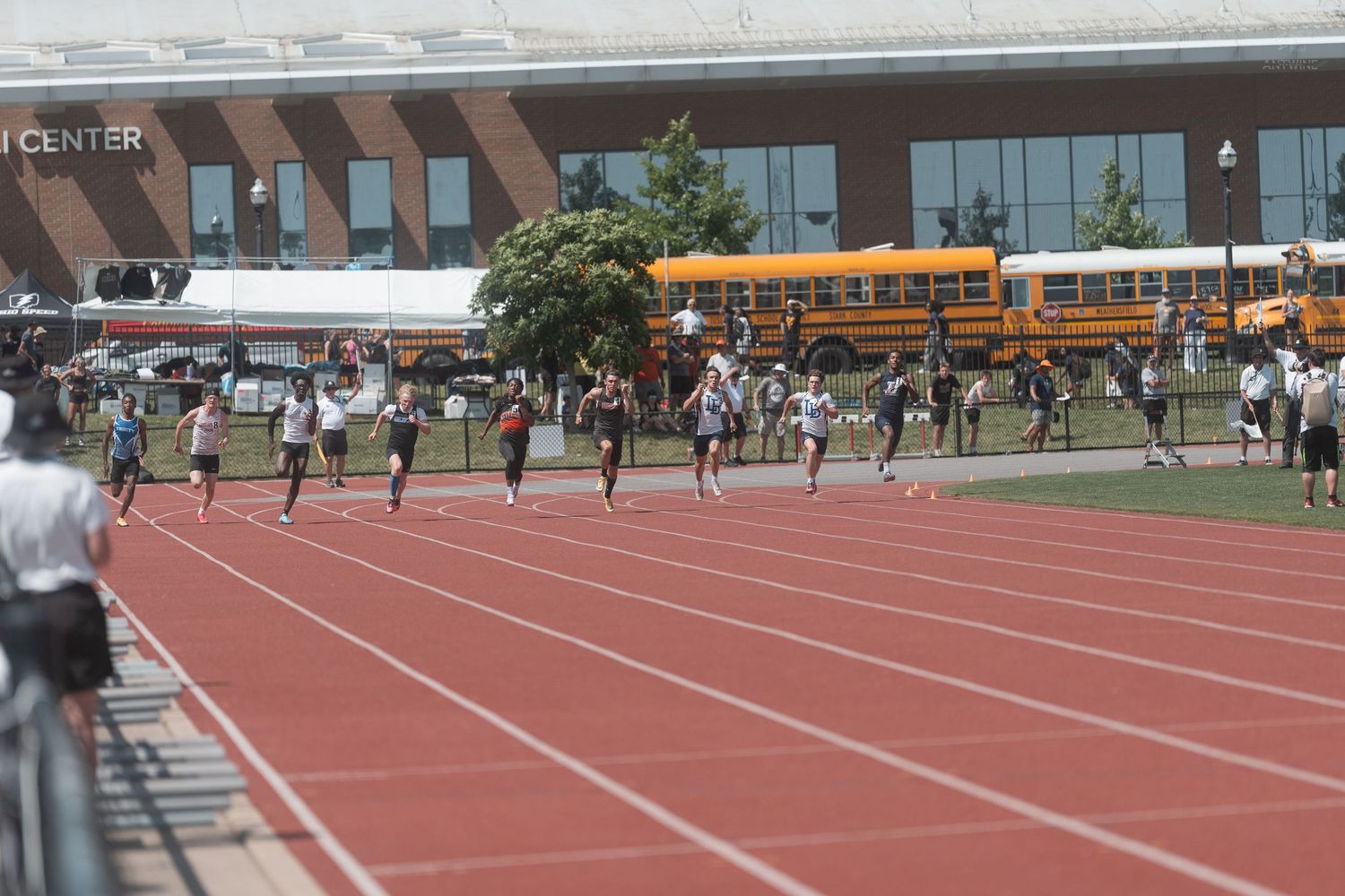 High school sprinters race down a red track at an outdoor track and field competition on a sunny day.