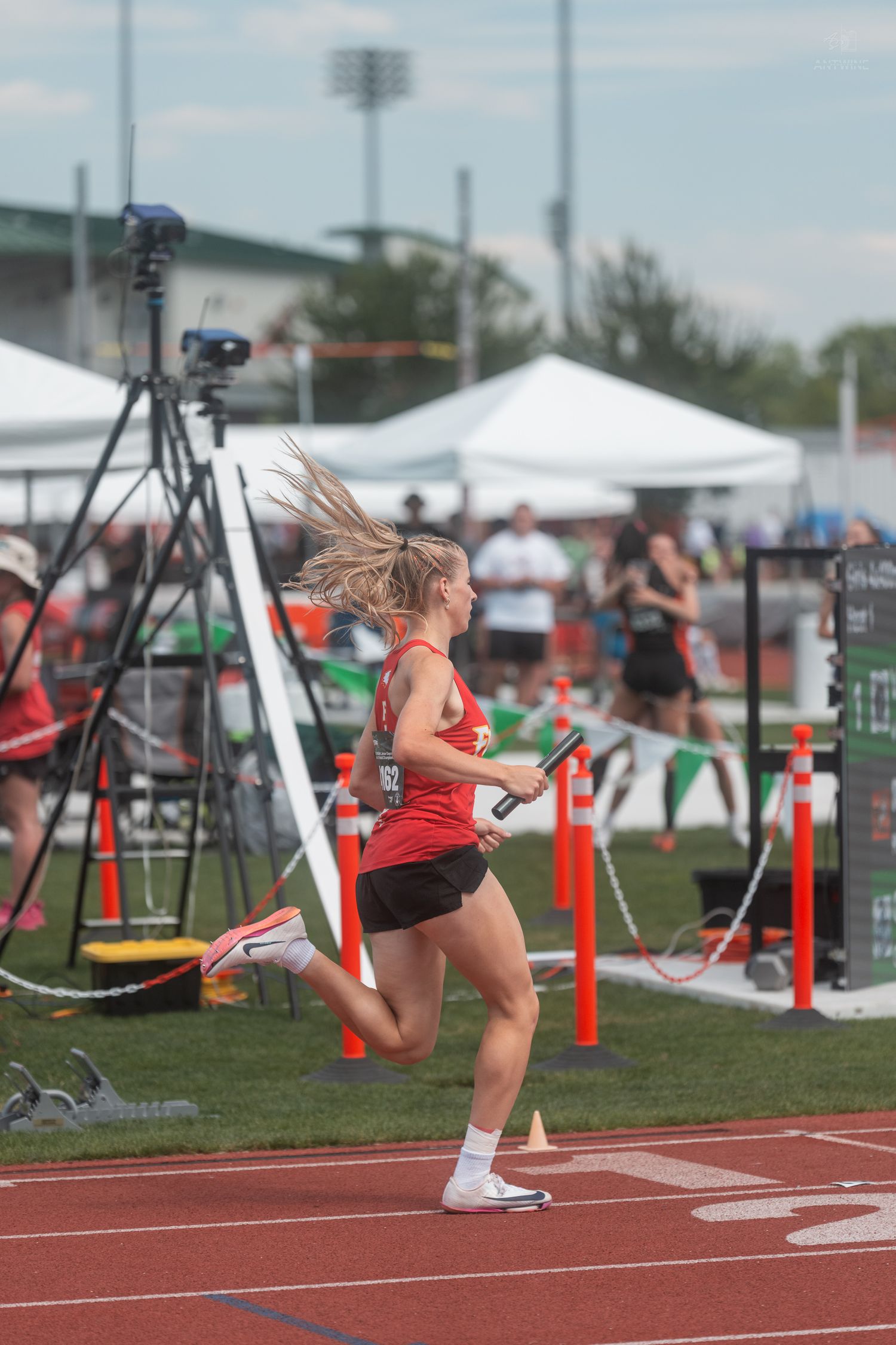 Female track athlete in red uniform runs with a relay baton, hair flying, during an outdoor track competition.