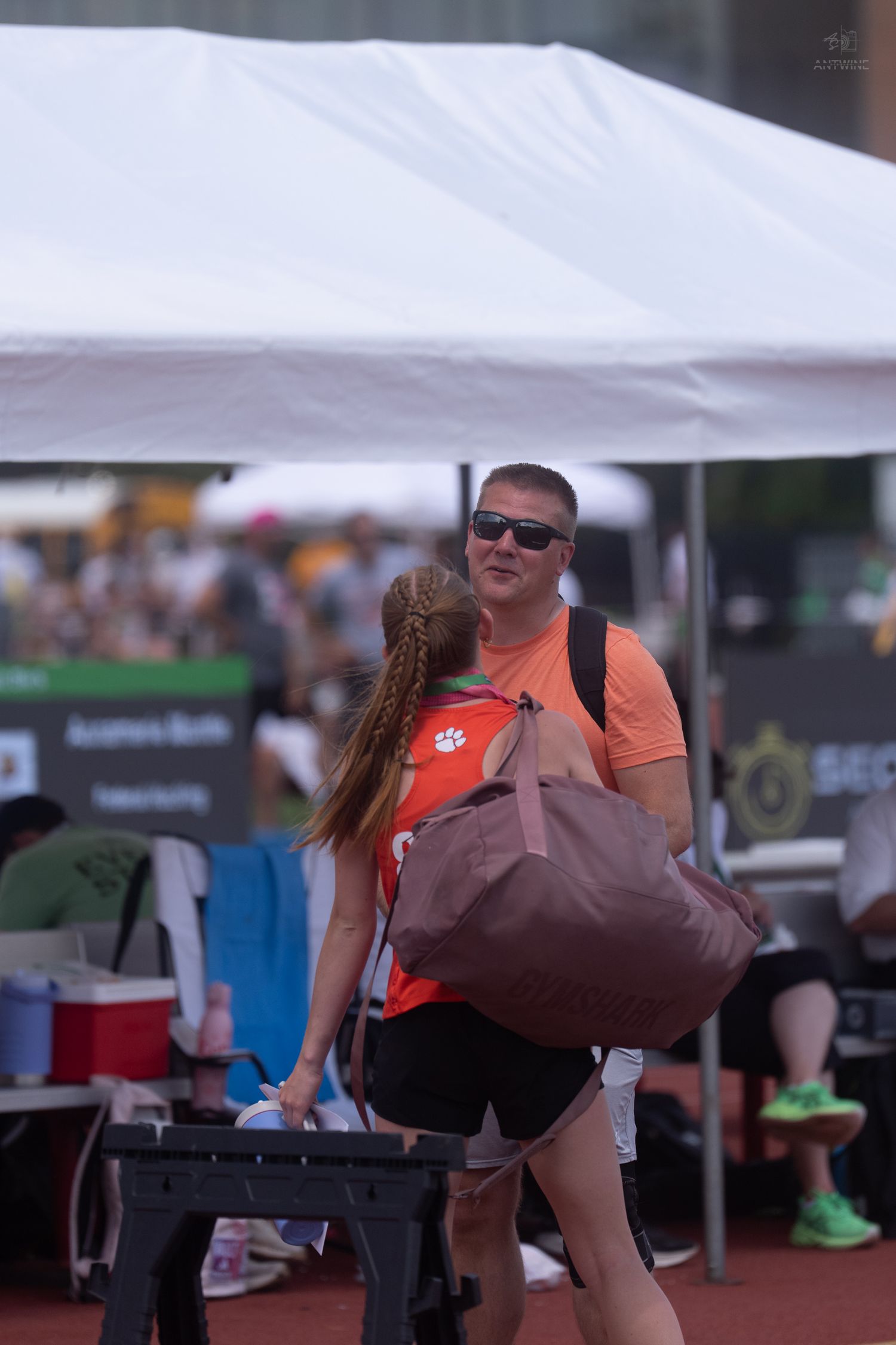 A man in an orange shirt carries a large bag while talking to a young female athlete at a track meet.