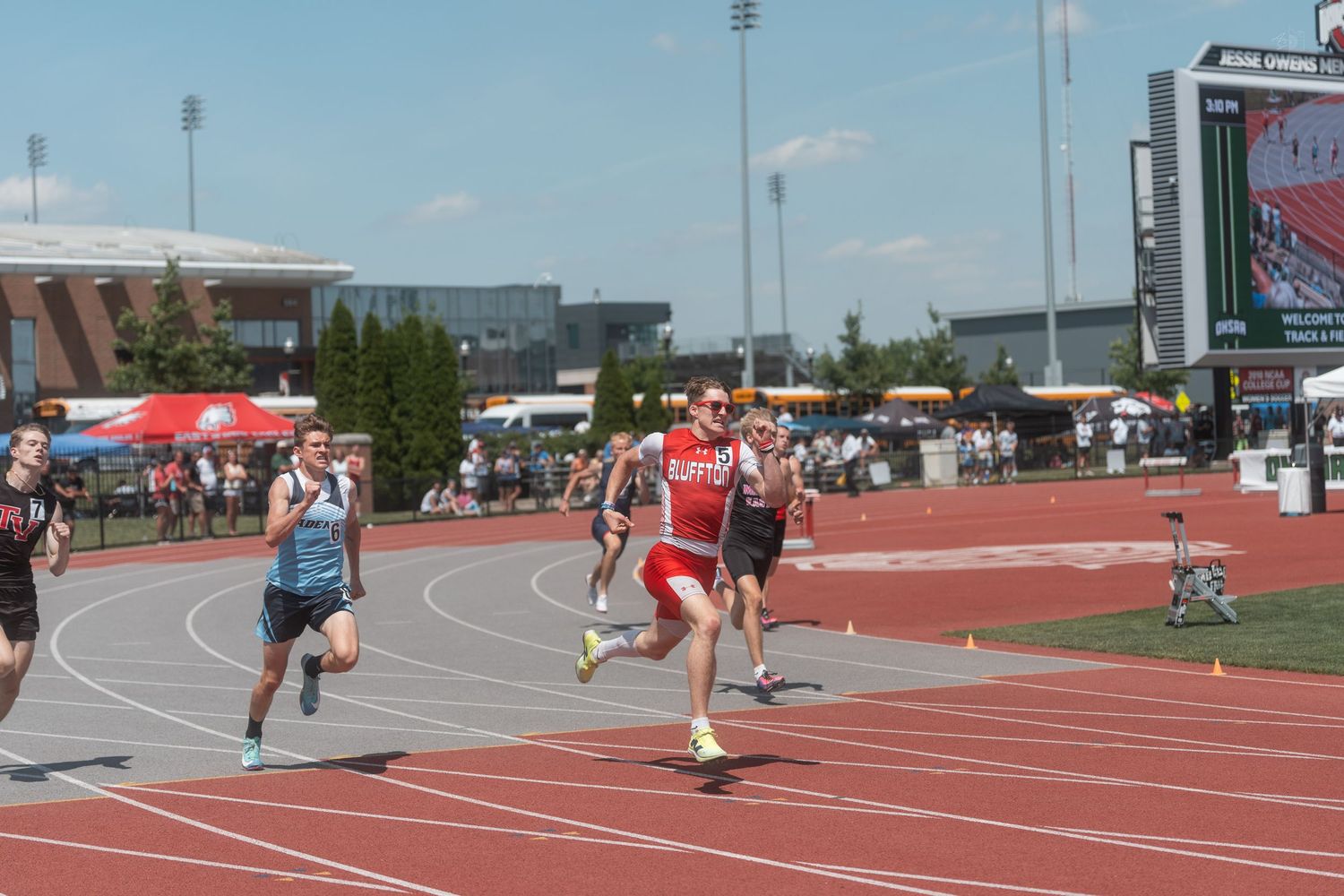Male runners competing in a sprint race on a red track at a sunny outdoor high school track meet.