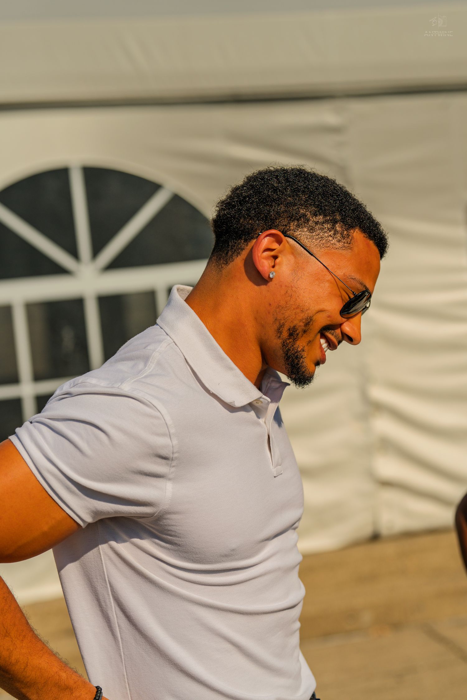 A smiling man with short curly hair wearing sunglasses and a white polo shirt outdoors near a white tent.