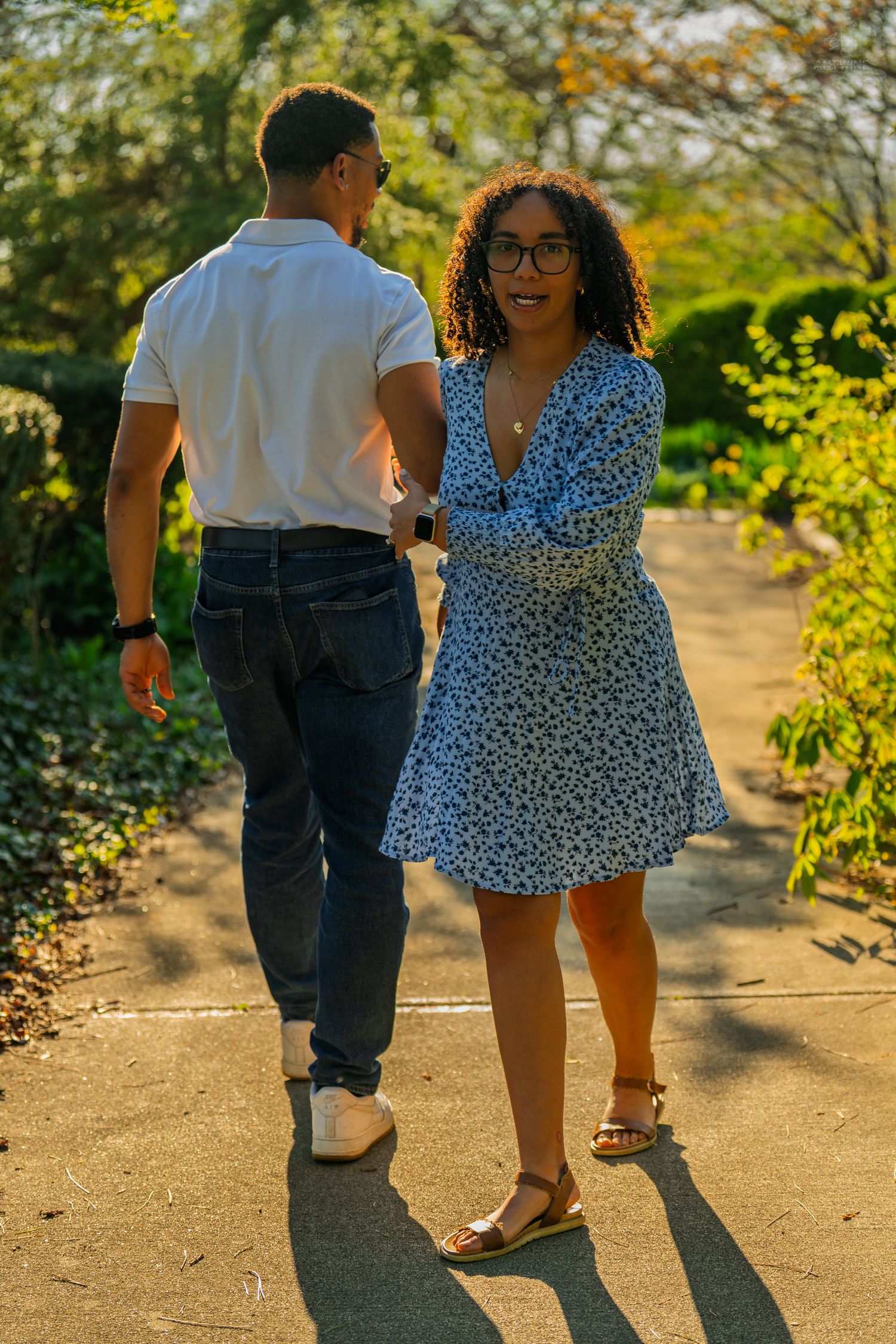A woman in a floral dress and glasses smiles back while walking with her partner on a sunny park path.