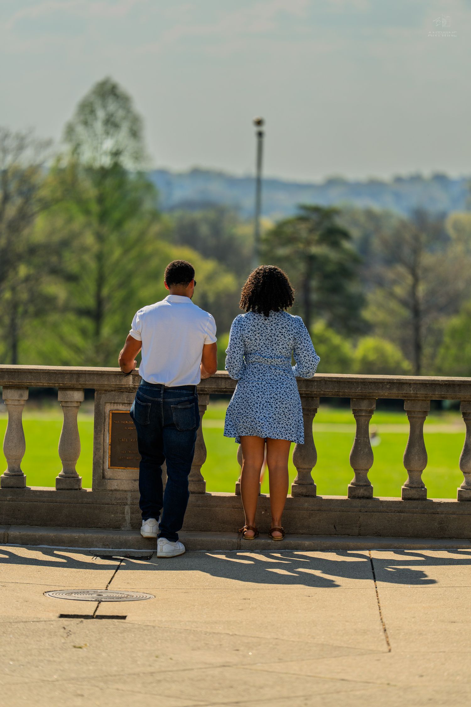 Couple standing at stone balustrade overlooking lush green park landscape on sunny day.