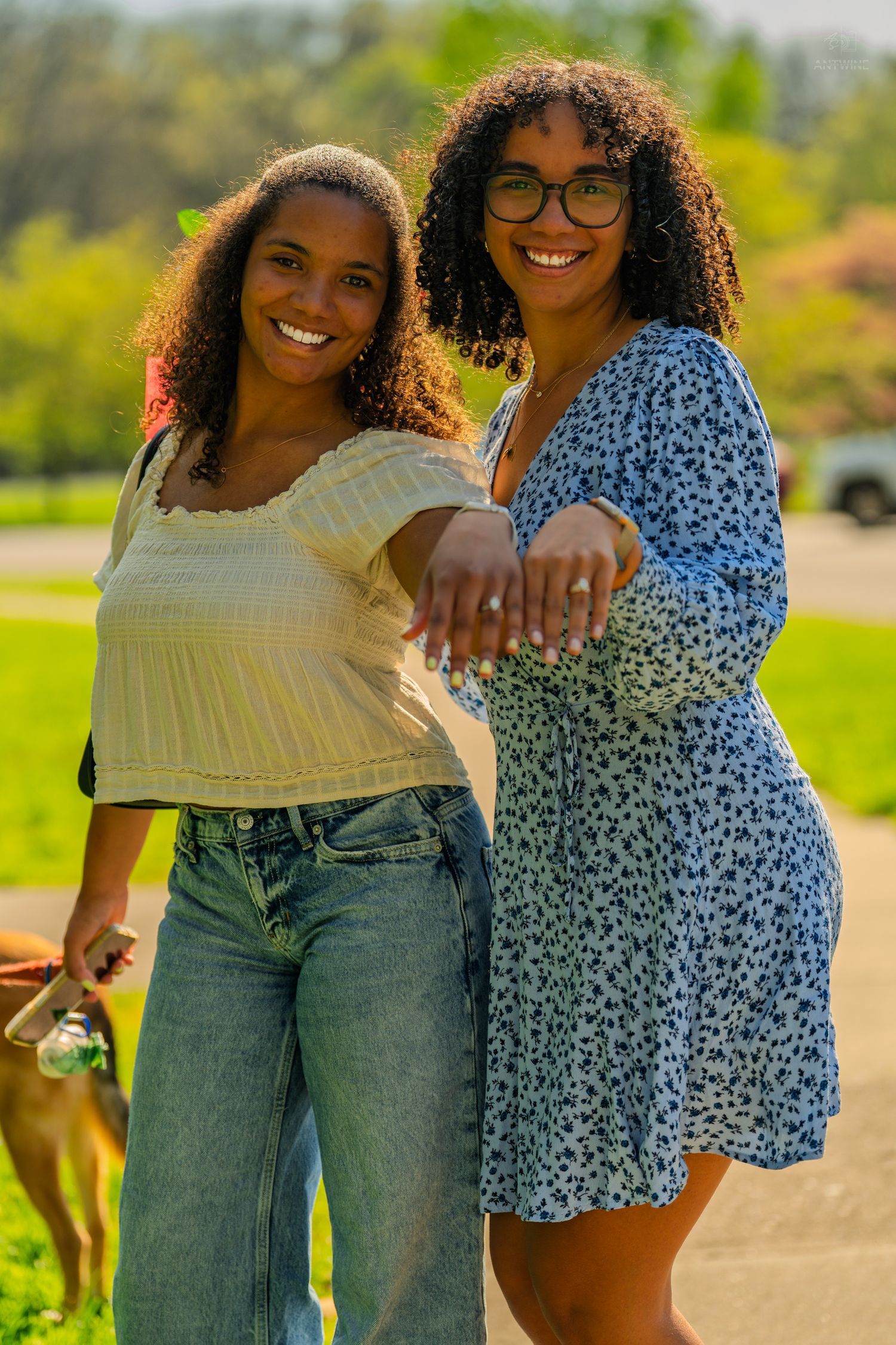 Two joyful women with curly hair smile and extend hands toward camera while posing in a sunny green park.