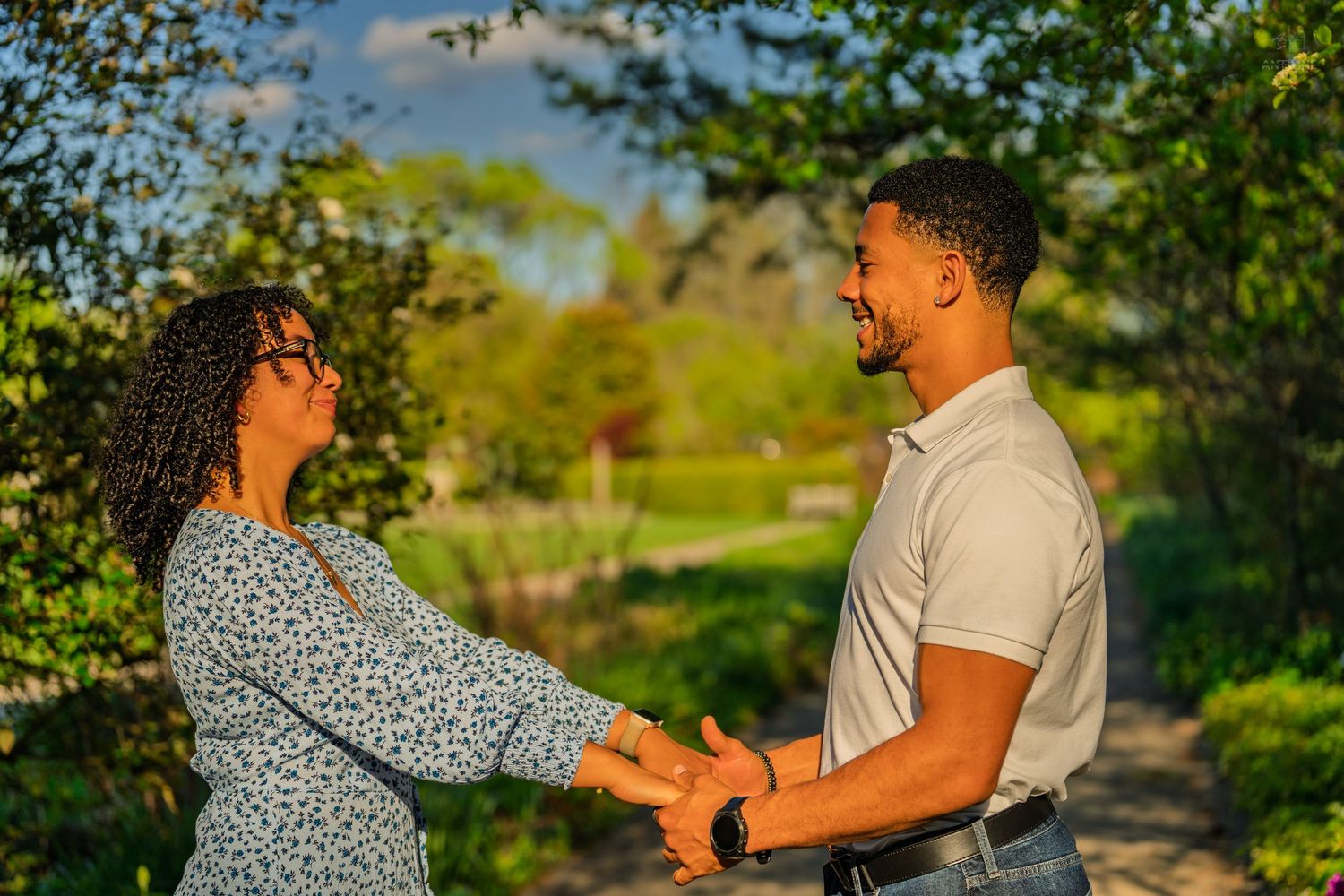 Happy couple holding hands and smiling at each other on a sunny park path surrounded by greenery.