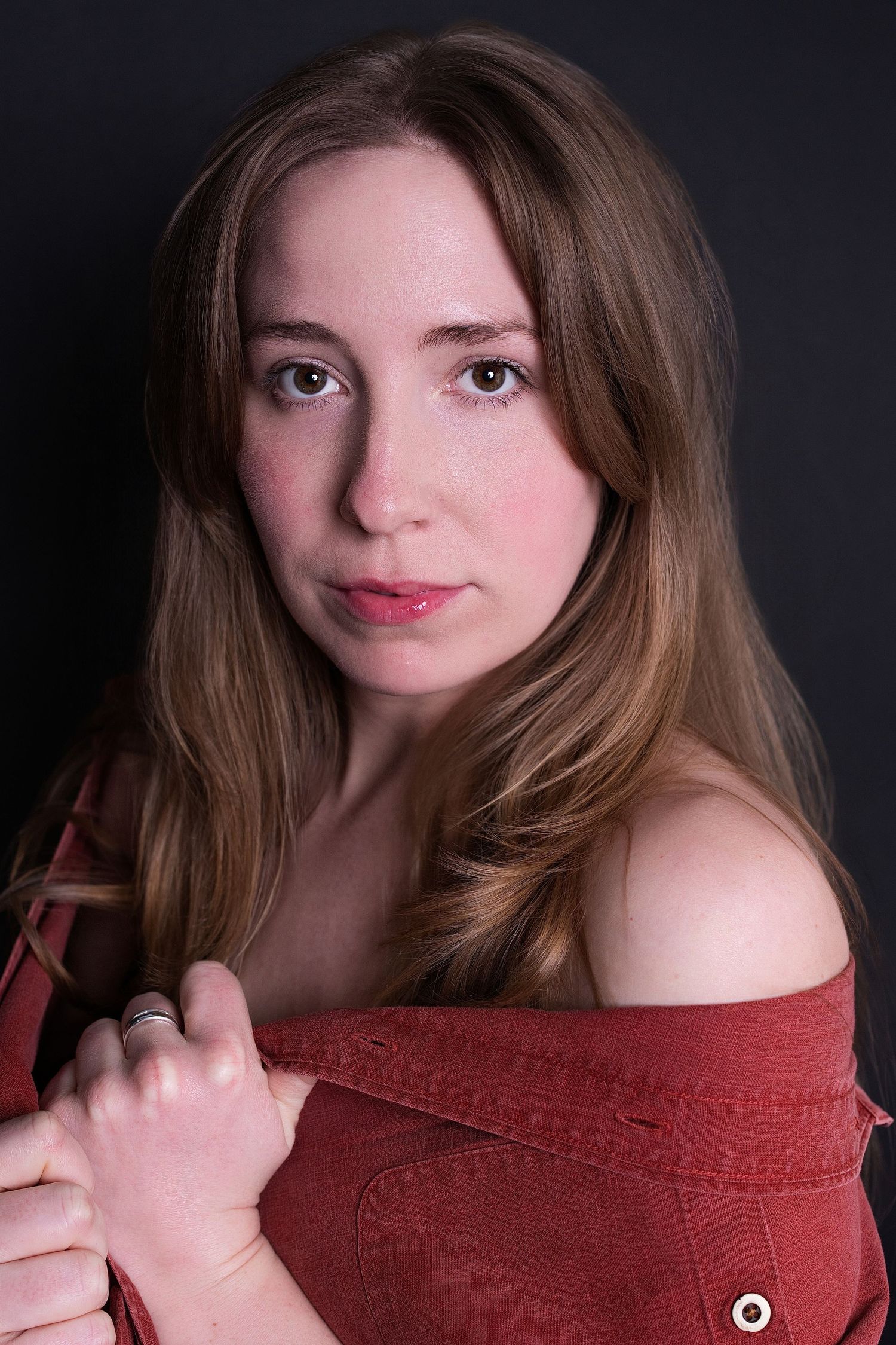 Young woman with long auburn hair posing in off-shoulder red jacket against dark studio background.