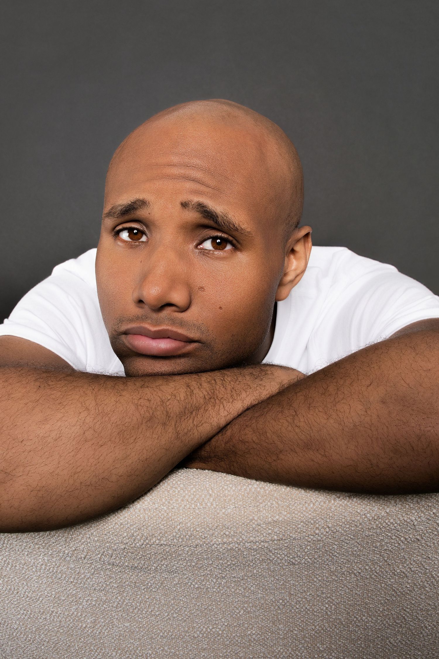 Bald Black man in white t-shirt with sad expression resting on crossed arms against gray background, color portrait.