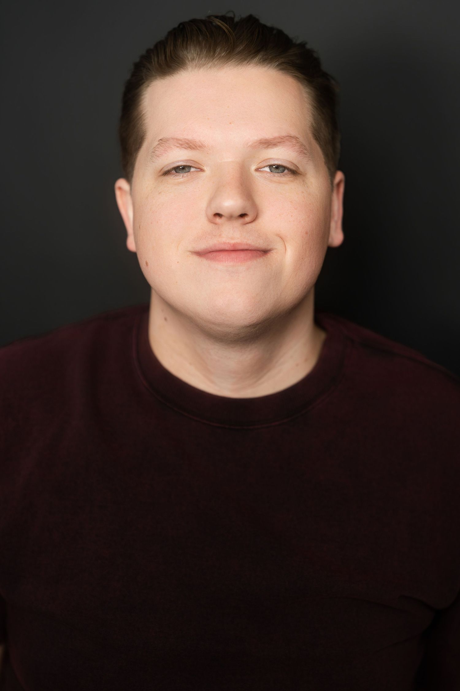 Headshot of smiling young man in maroon crewneck sweater against dark gray background.