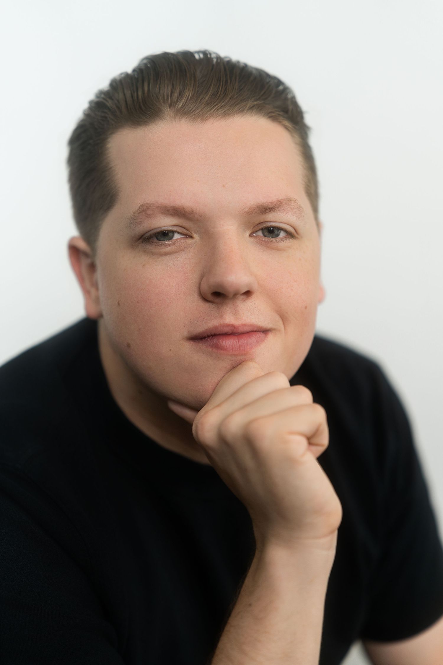 Young man in black t-shirt resting chin on fist with slight smile against white background.