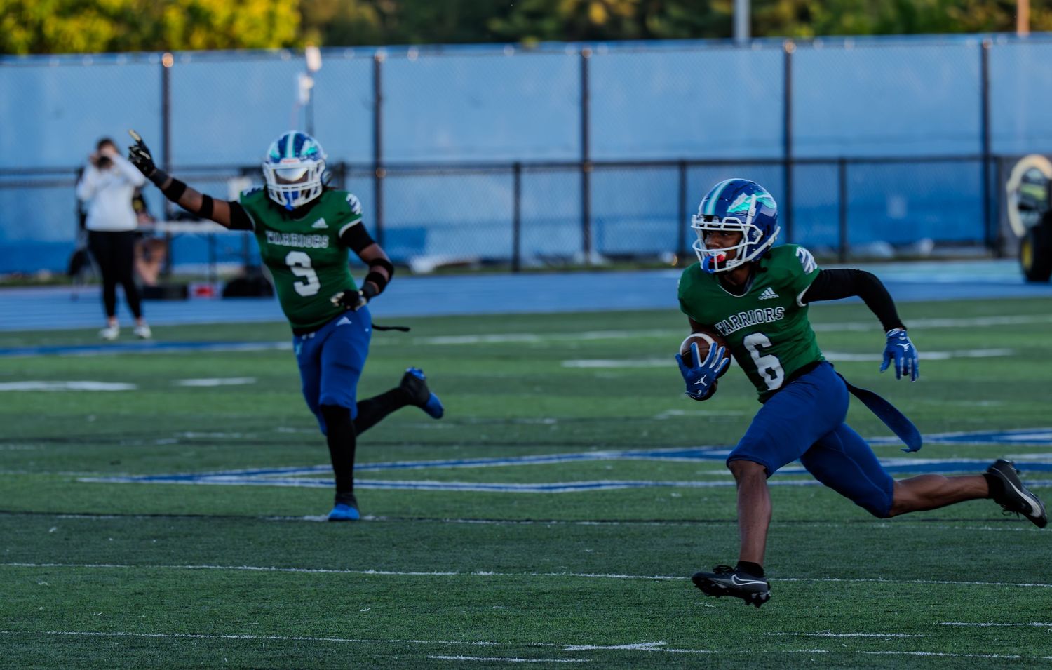 Two football players in blue and green uniforms run downfield during a daytime game.