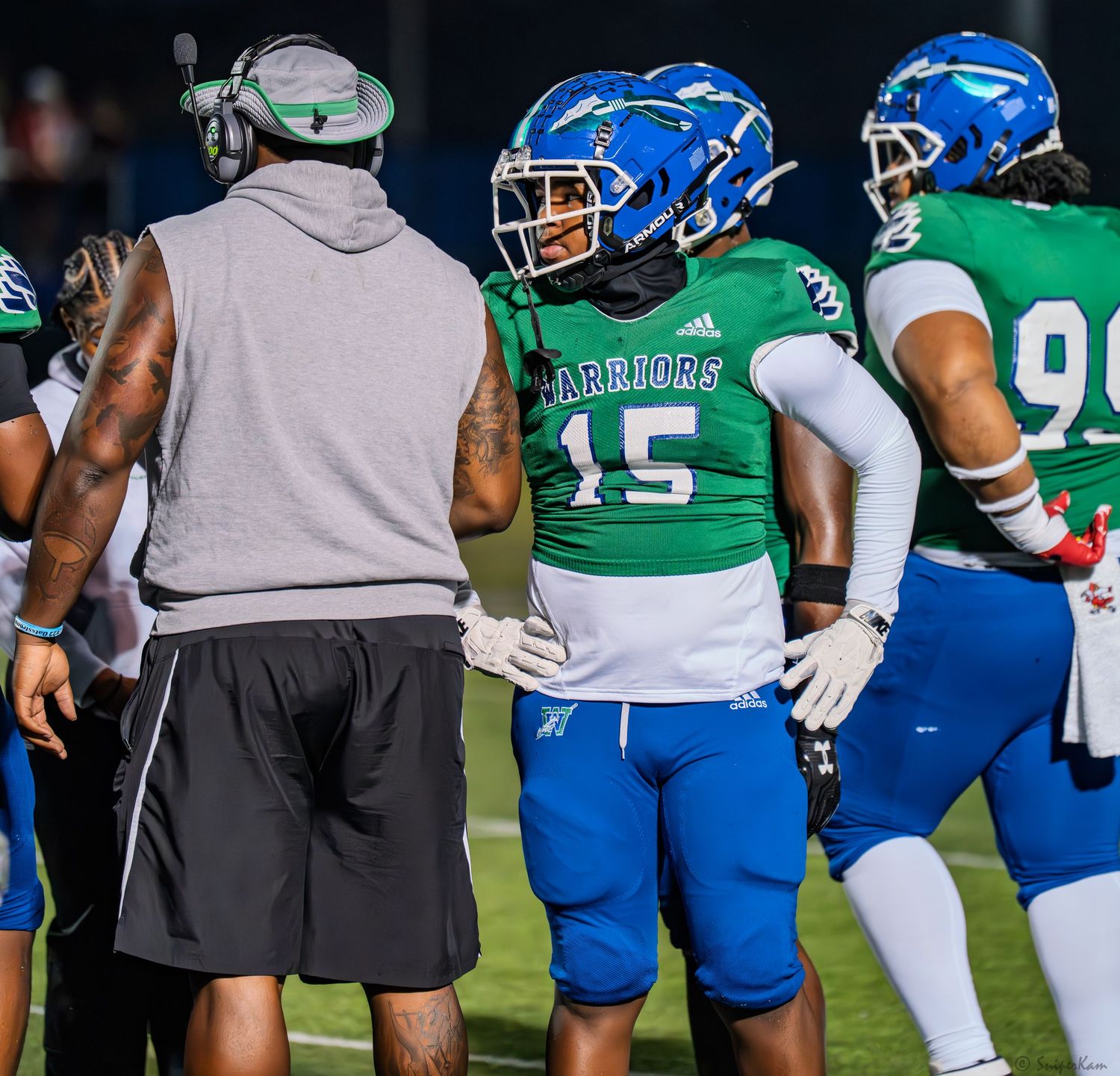 Football player number 15 in green Warriors uniform talks with coach on sideline during game.