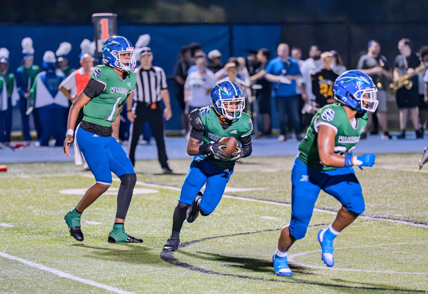 Three high school football players in green and blue uniforms run with the ball during a night game.