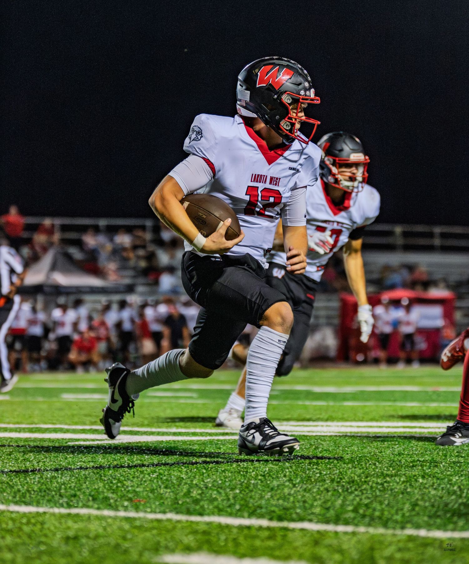 High school quarterback in white uniform runs with football along sideline during night game.