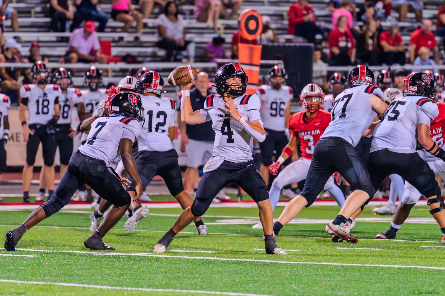 High school quarterback #4 in red uniform prepares to throw a pass behind his offensive line during a night football game.