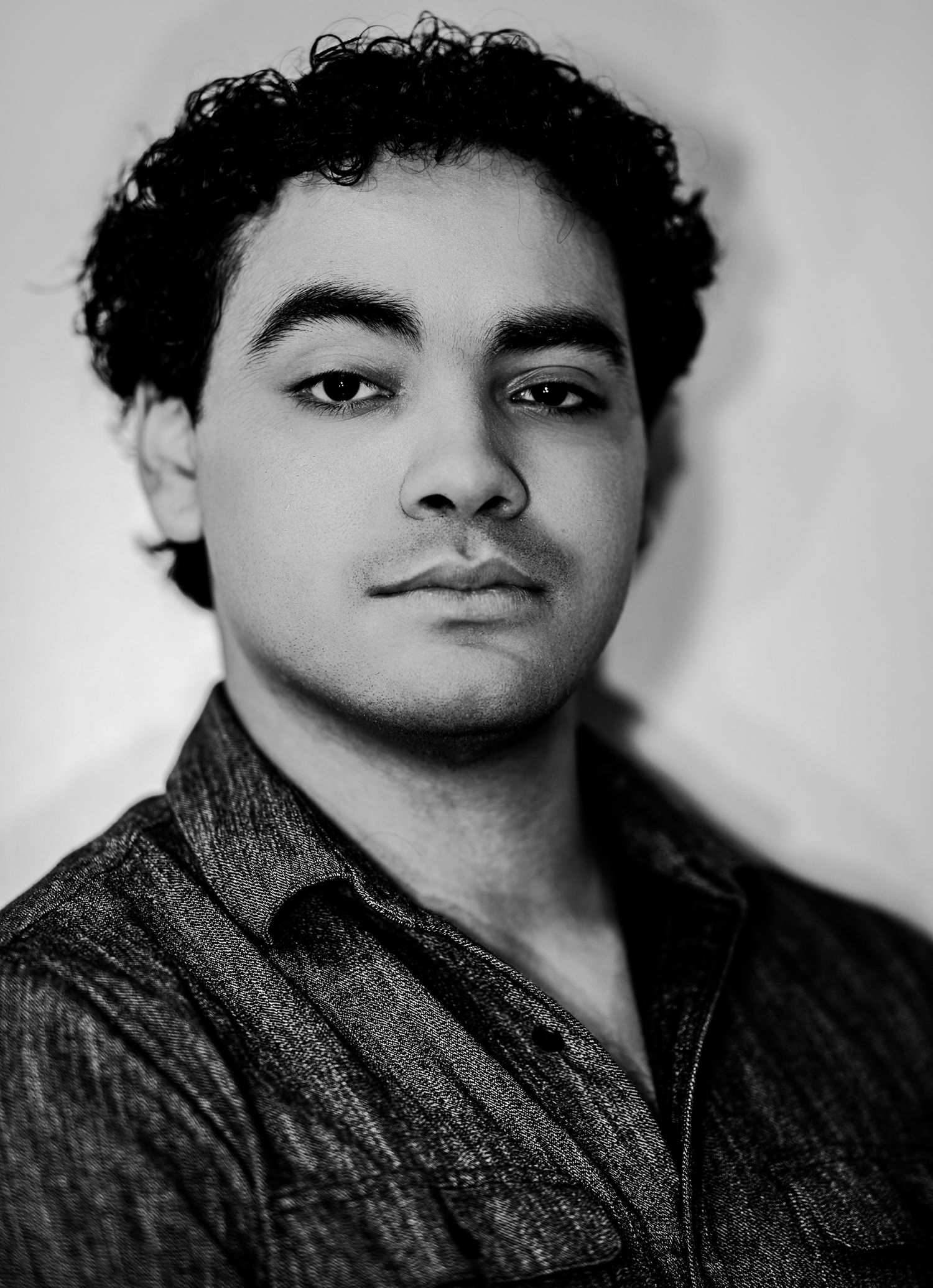 Young man with curly dark hair in a dark shirt with a serious expression in a black and white portrait.