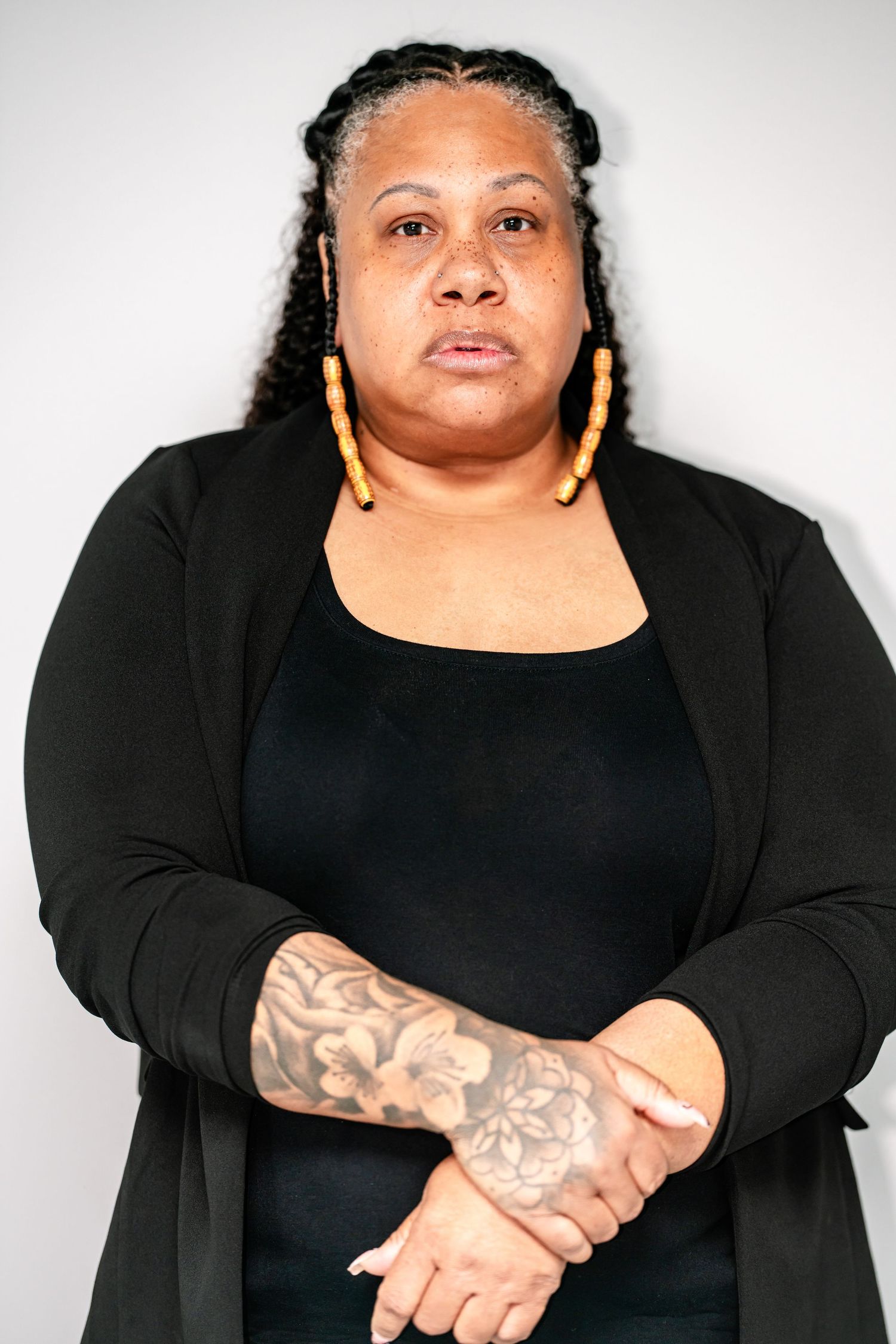 Black woman in black outfit with floral tattoo on arm and yellow hoop earrings, serious expression.