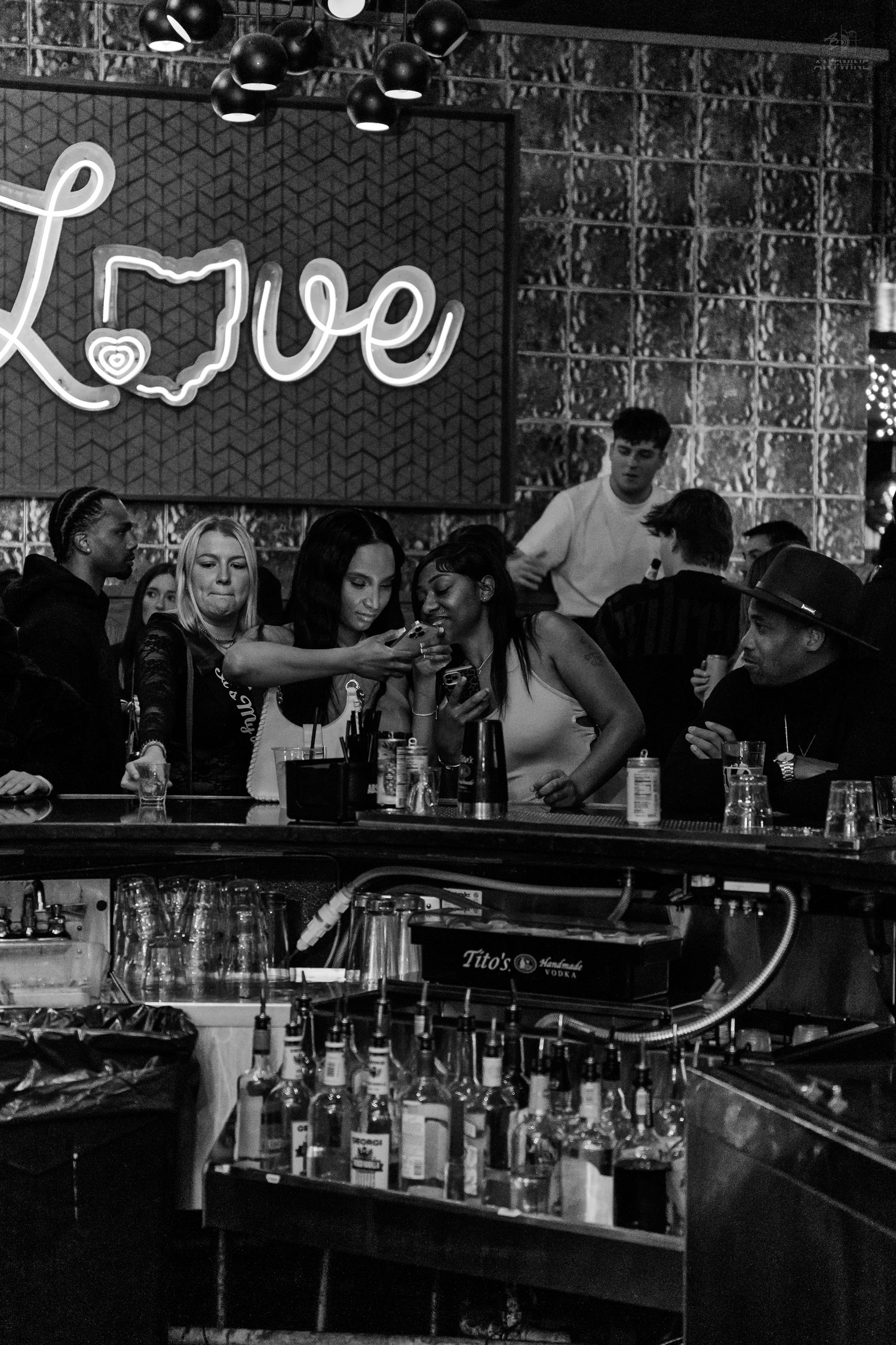 Black and white photo of women taking a selfie at a bar with a neon Love sign.