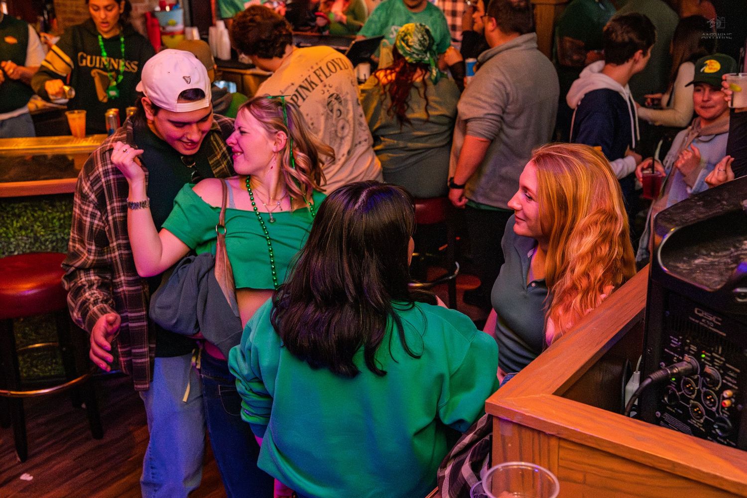 Young women in green tops socializing and laughing at a St. Patrick's Day bar event.