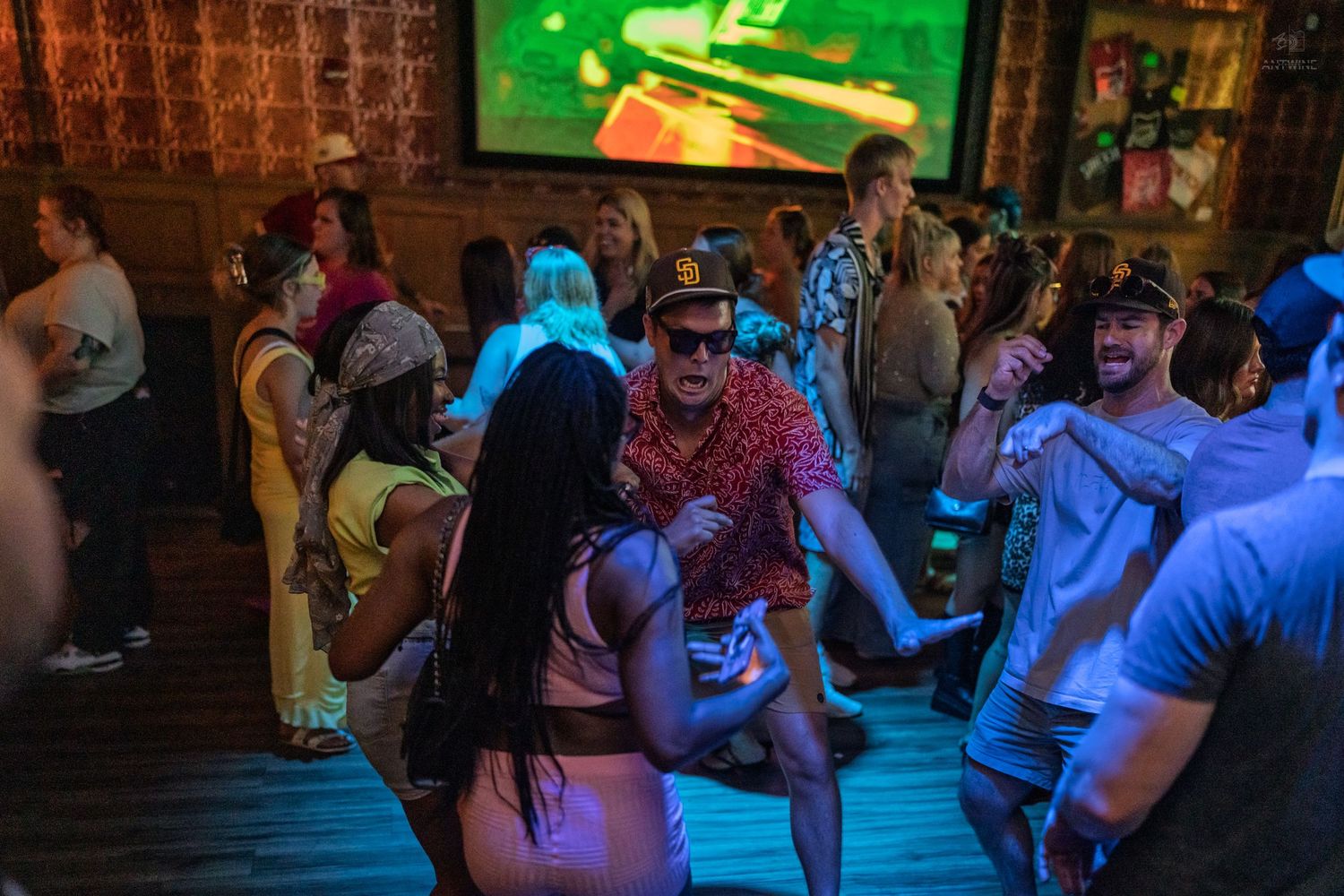 Energetic crowd dancing inside a bar with a large projection screen and blue stage lighting.