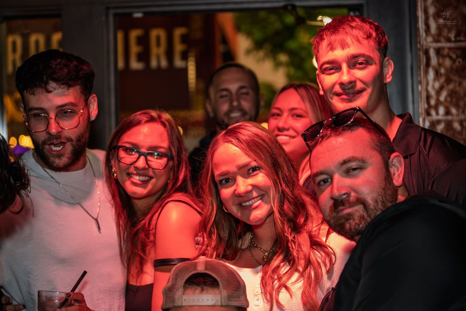 Group of six friends smiling together under red ambient lighting at a nightlife venue.