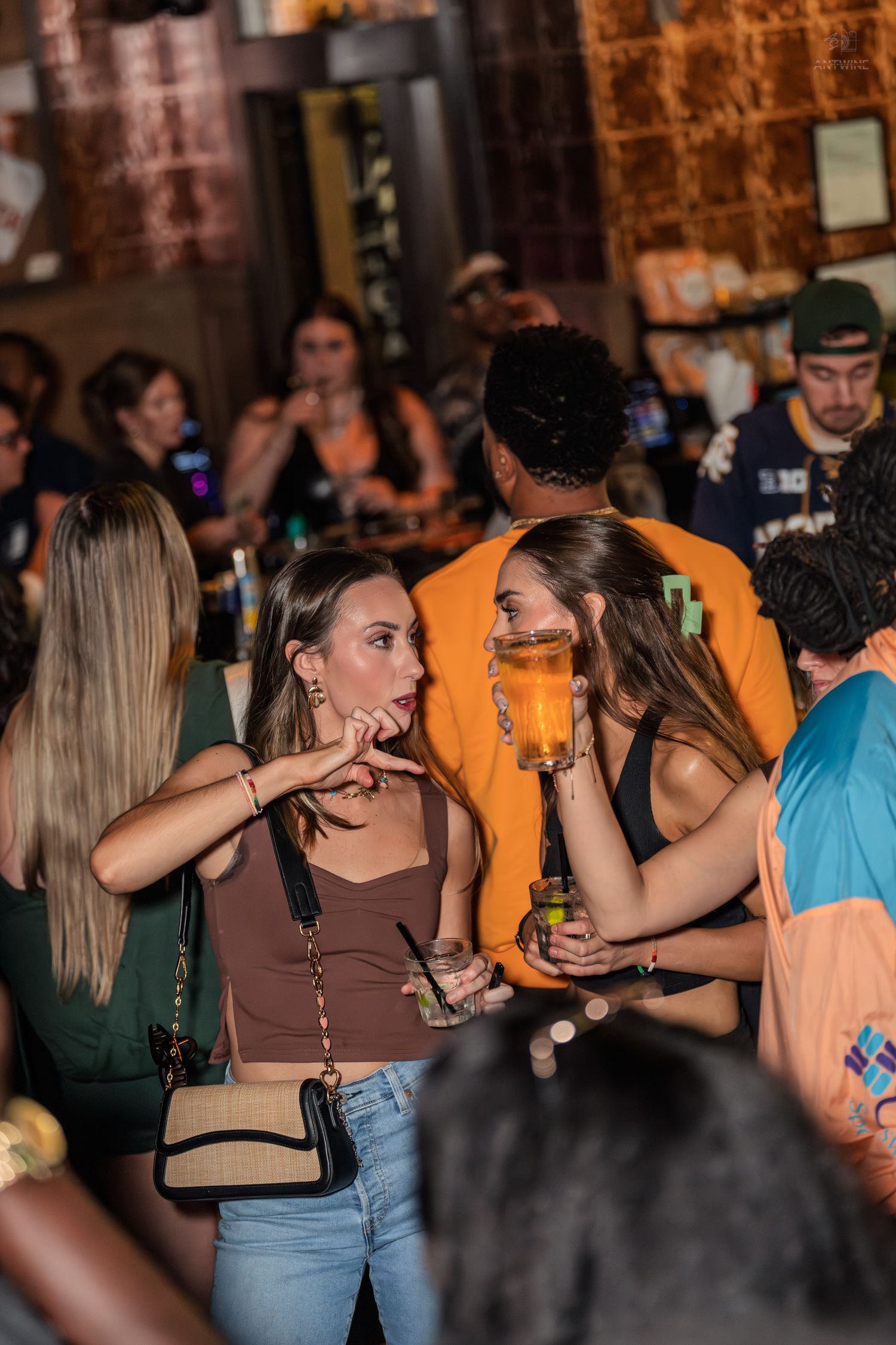 Two women talking and holding drinks in crowded bar venue.