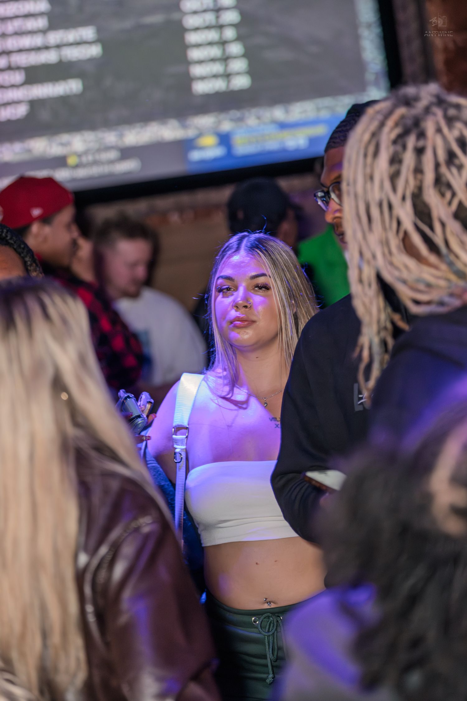 Young blonde woman in a white crop top standing in a crowded nightclub with purple lighting.