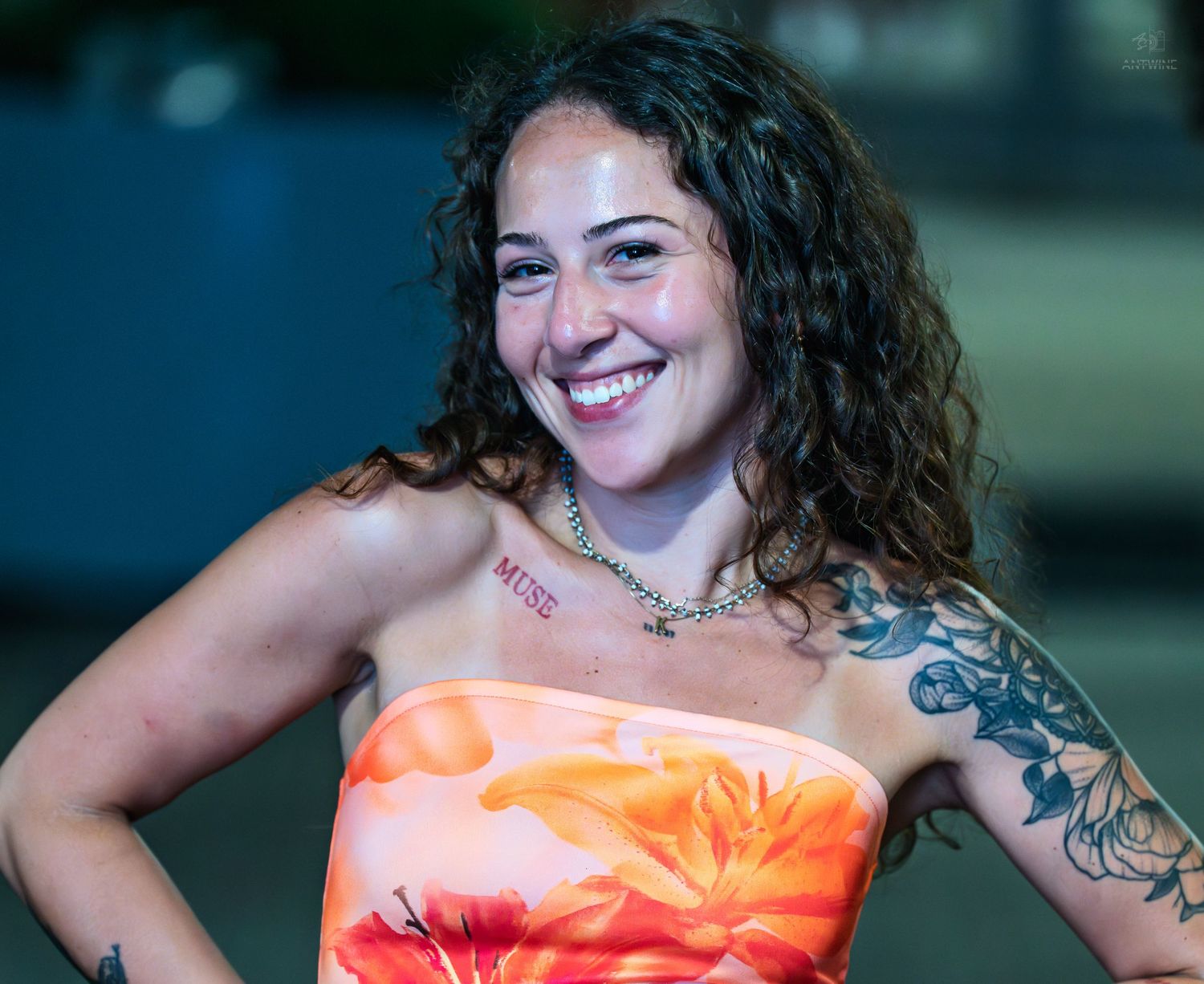 A happy woman with curly hair and tattoos wearing an orange floral strapless top smiles at an outdoor event.
