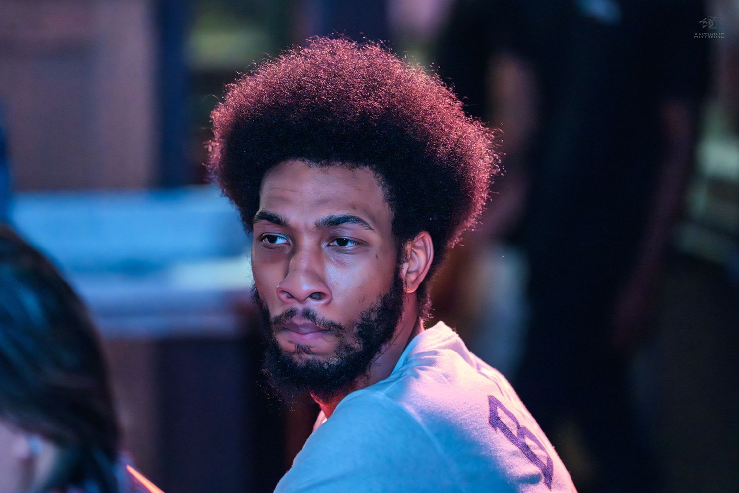A young Black man with a large afro hairstyle sits in a dimly lit venue, looking to the side.