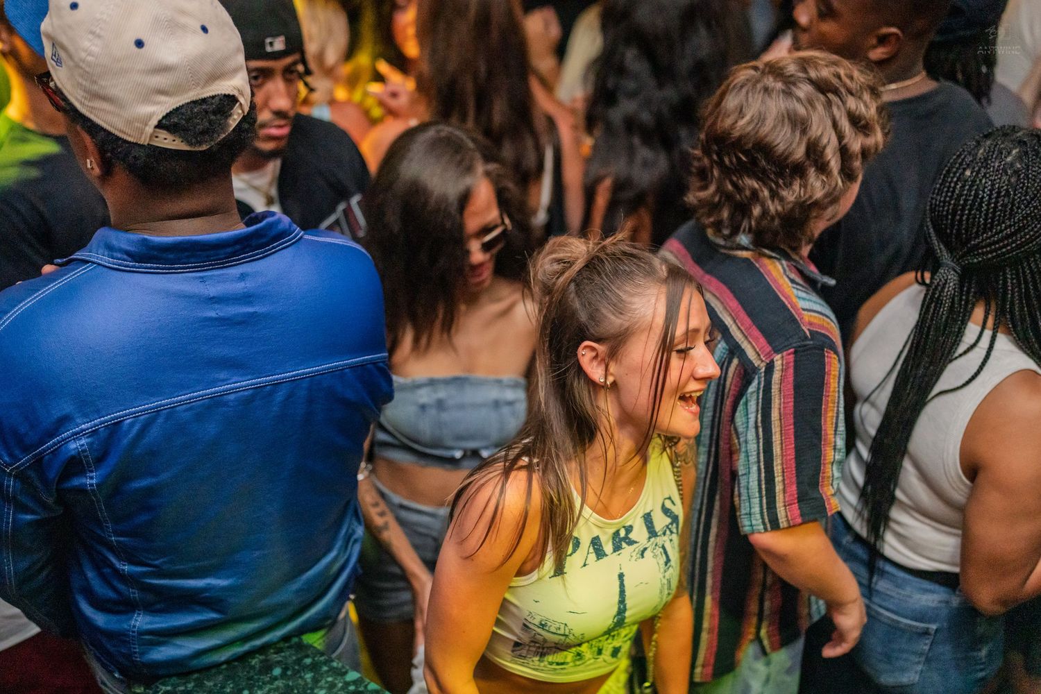 Young woman in yellow top dancing joyfully in a dense nightclub crowd with colorful lighting.