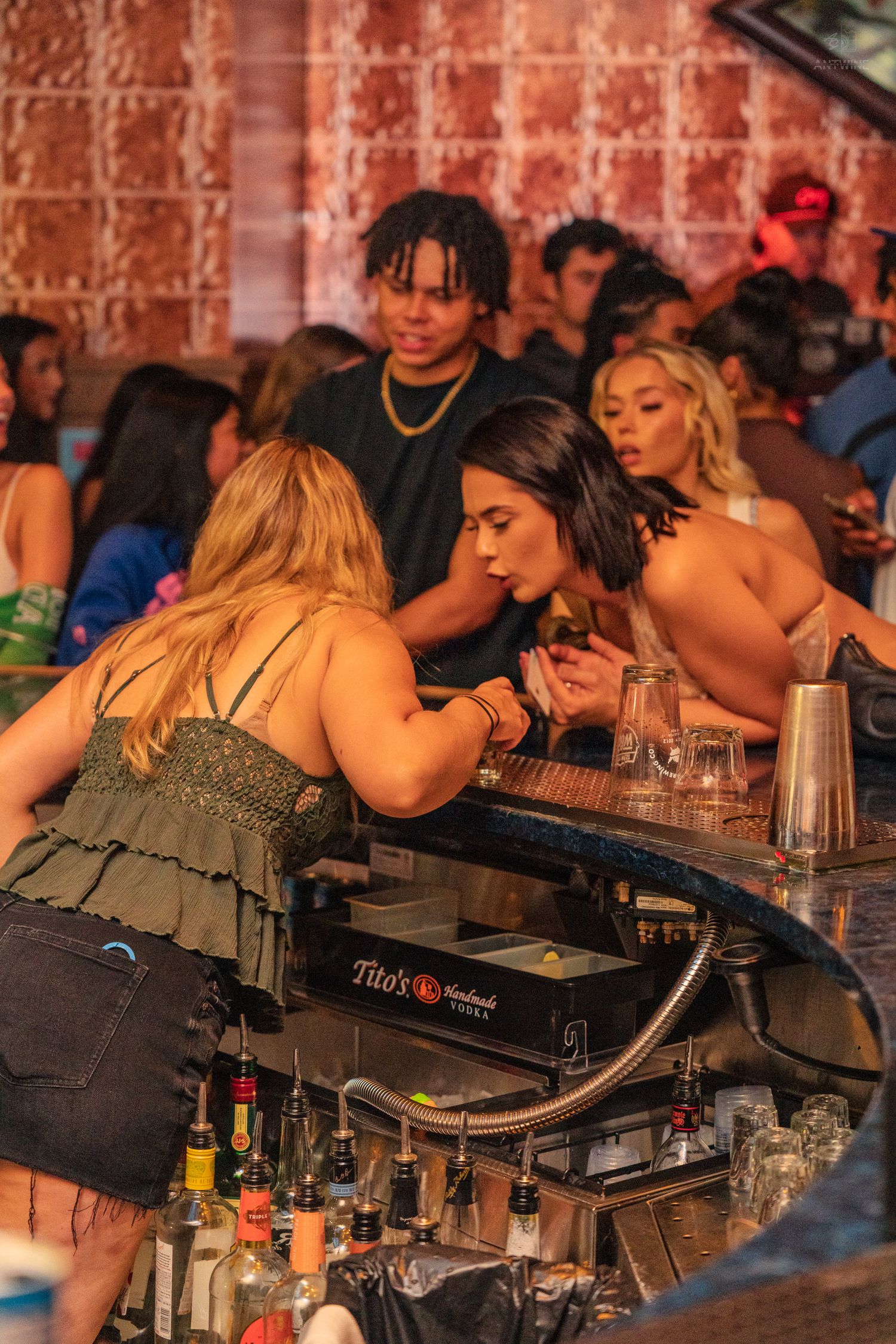 Women leaning over a bar counter at a nightclub with Tito's vodka display and bottles.