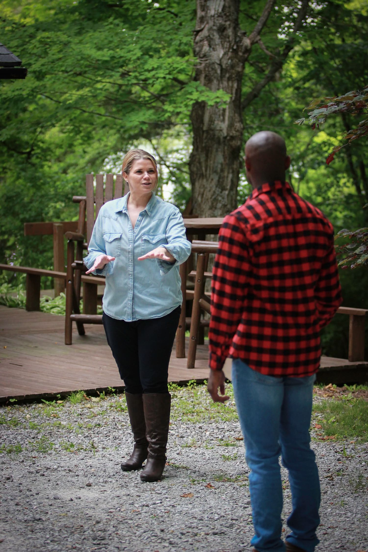 A blonde woman in a denim shirt gestures while speaking to a man in a red plaid shirt outdoors in a wooded area.