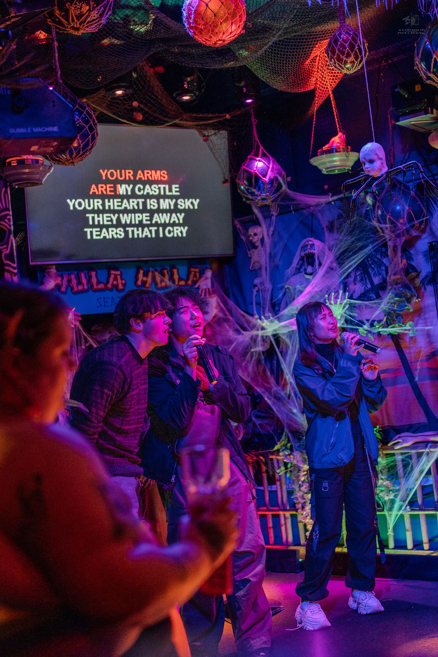 Group singing karaoke at a Halloween-decorated bar with cobwebs and lyrics displayed on screen.