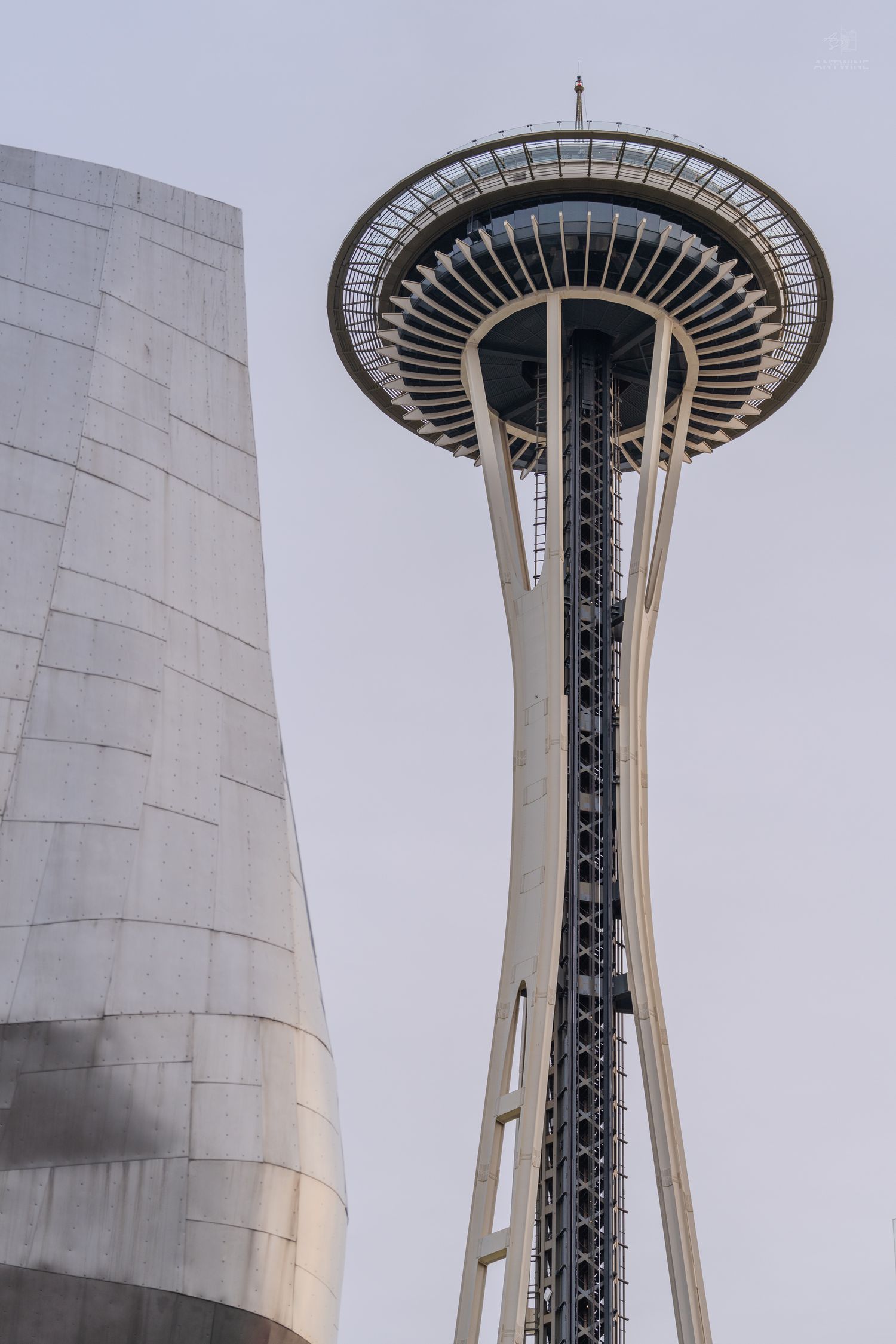 The iconic Space Needle tower in Seattle, Washington, rising against a gray overcast sky with a modern building visible to the left.