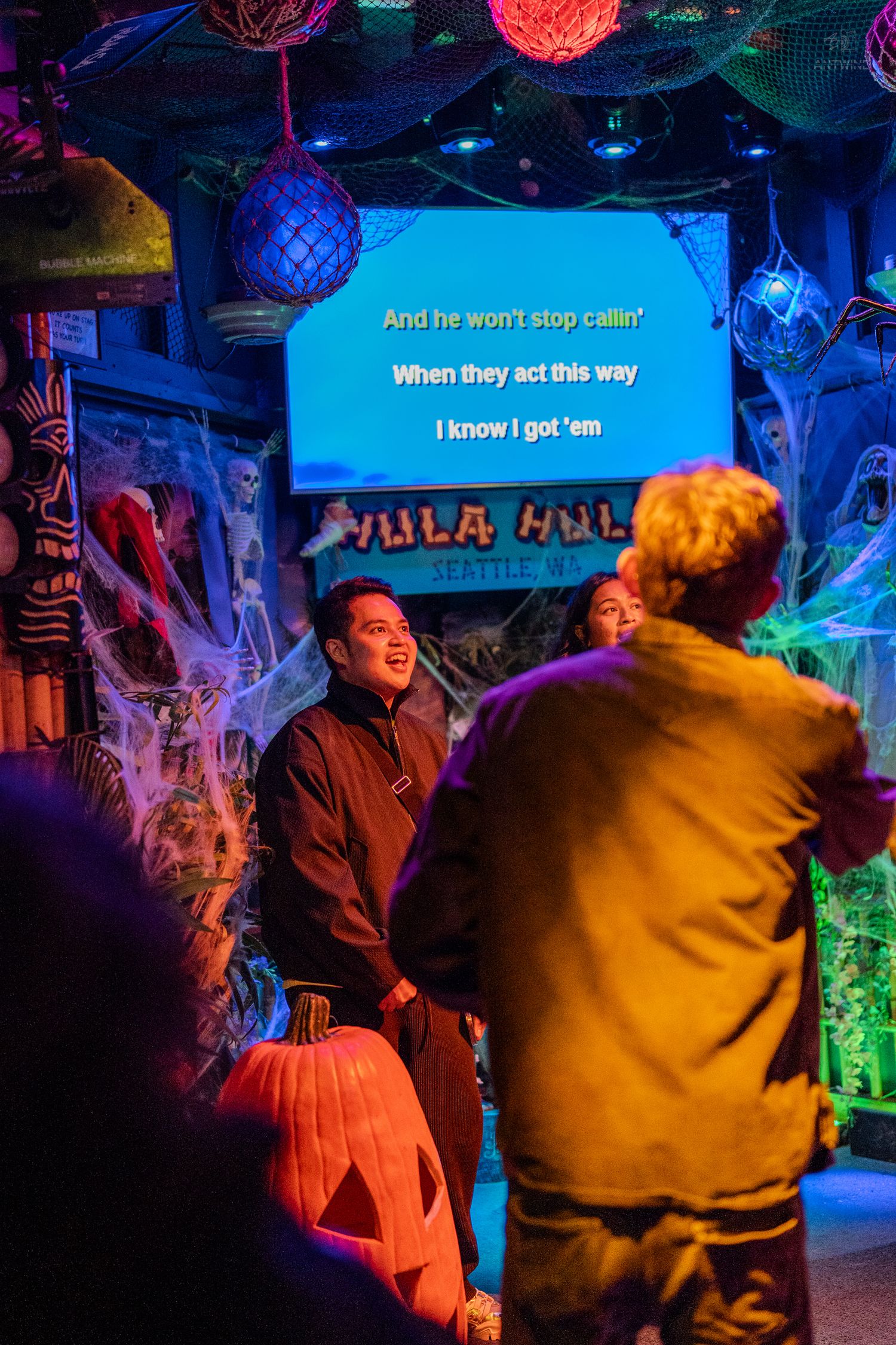 Friends enjoying karaoke night at Hula Hula Seattle bar with Halloween decorations and blue screen lyrics.