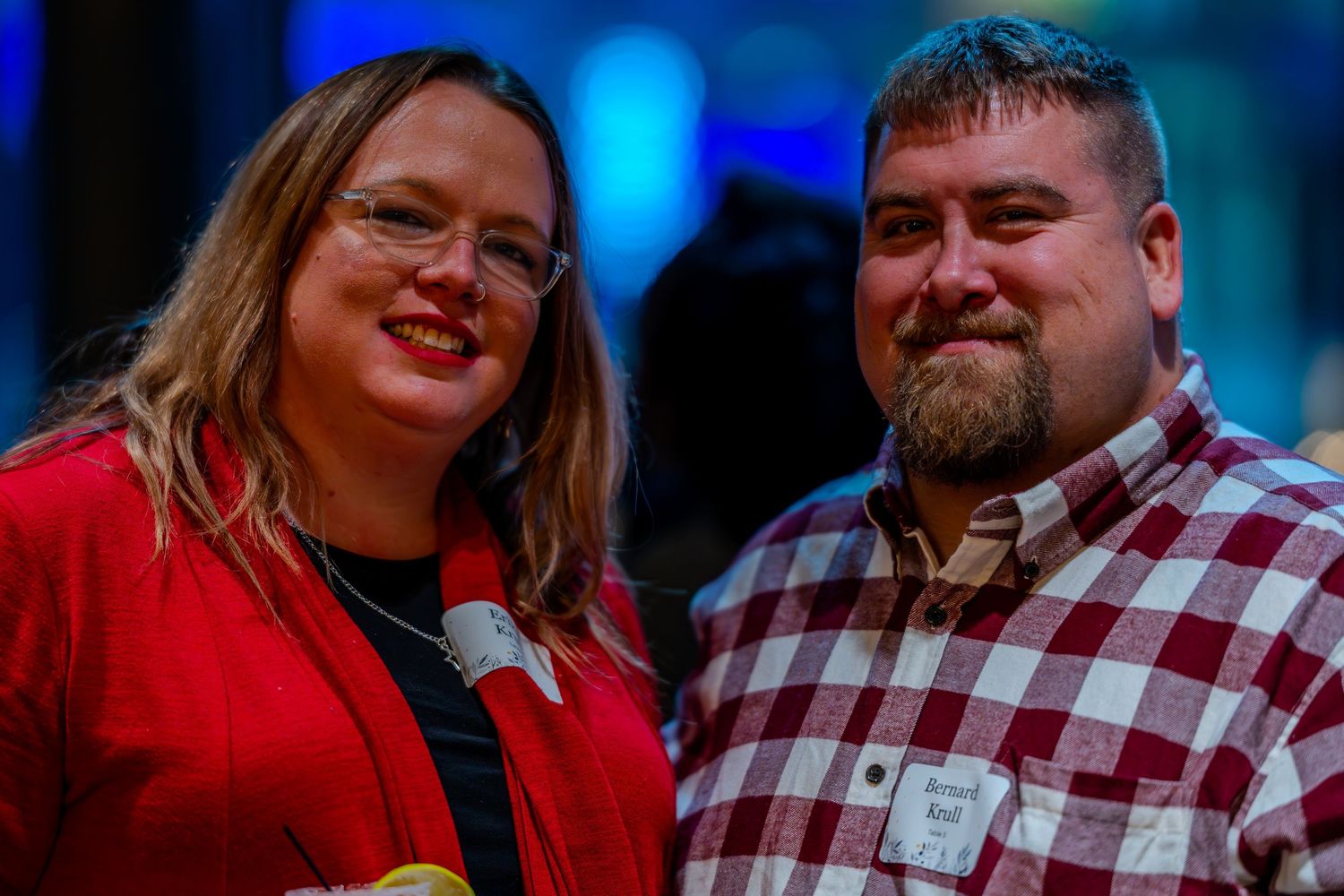 A smiling man and woman pose together at an indoor event, woman in red jacket, man in plaid shirt.