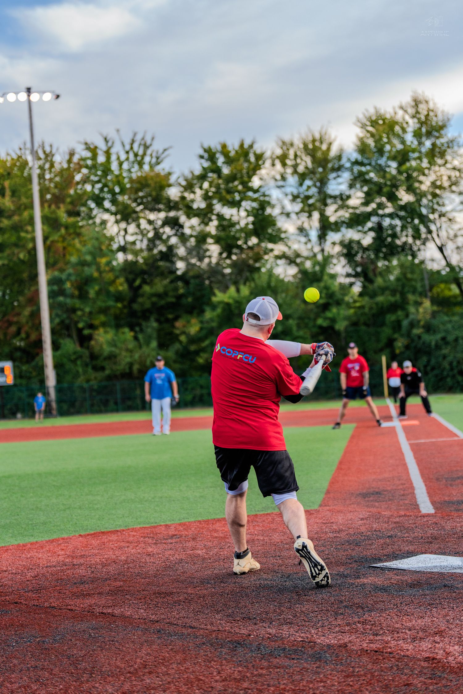 A player in a red jersey swings at a yellow softball during an evening recreational baseball game on a red turf field.