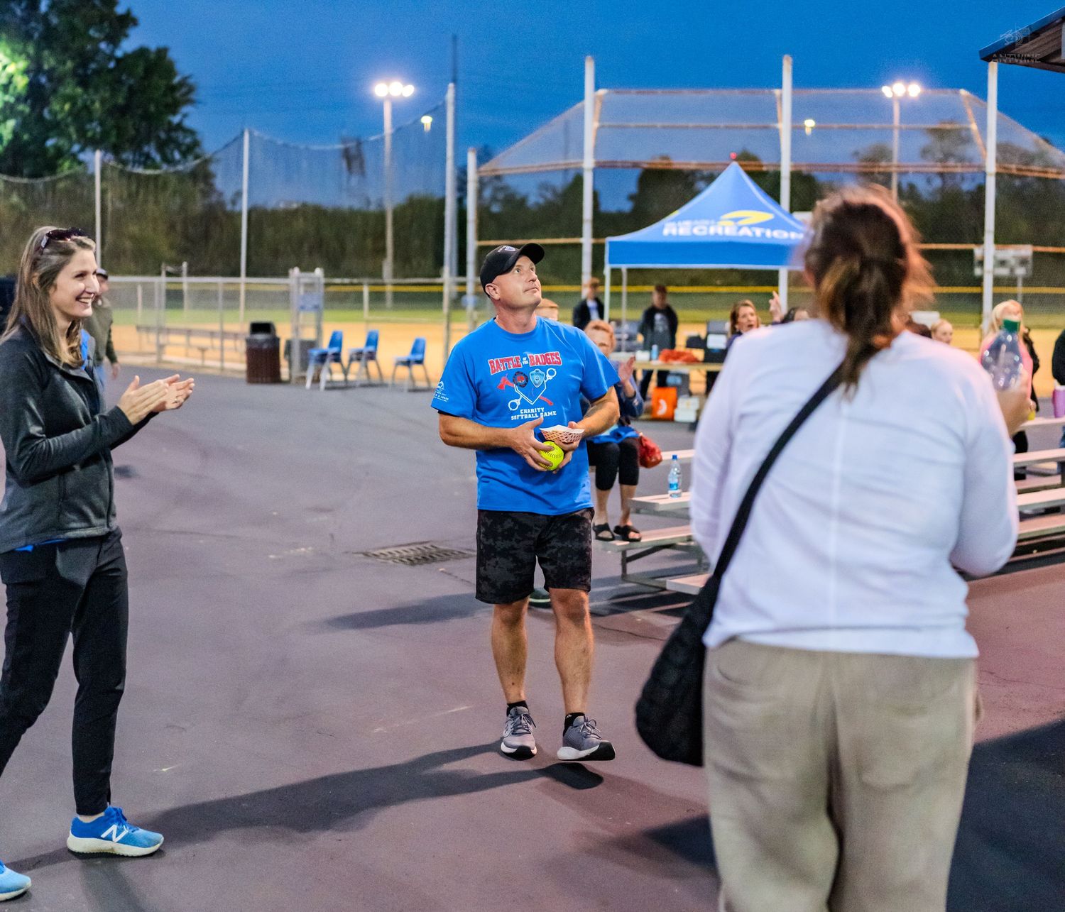 A man tosses a softball at an outdoor sports complex at dusk while two women watch nearby under bright field lights.