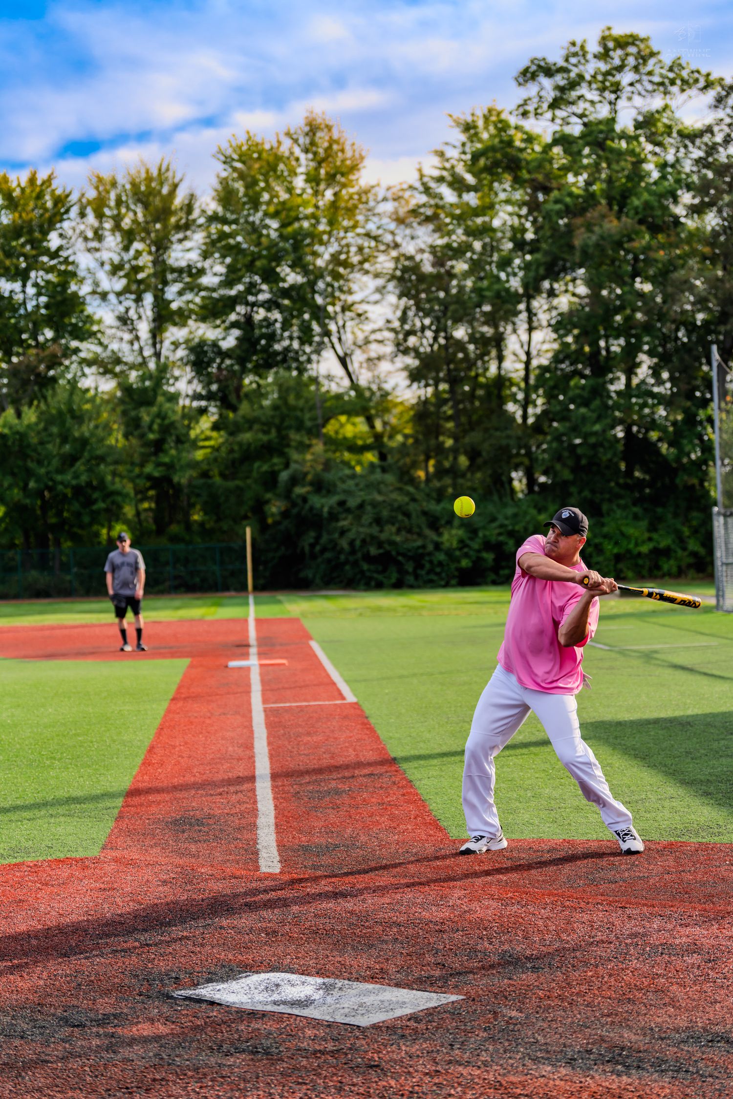 Softball batter in pink shirt swinging at yellow ball on red turf field with fielder in background.