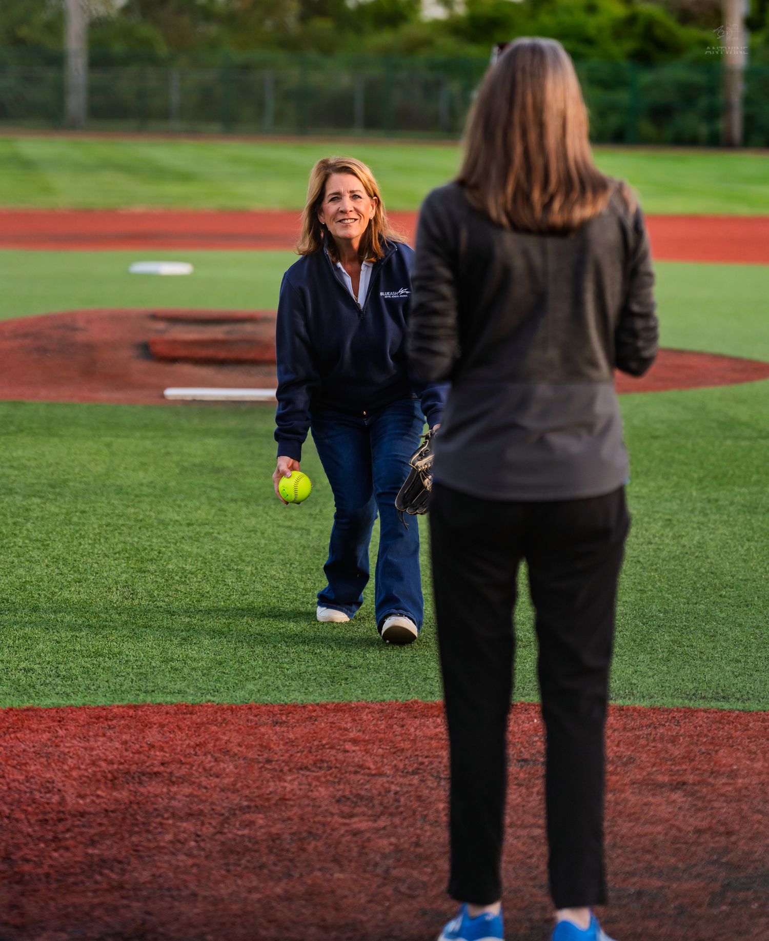 Woman in navy hoodie prepares to throw a yellow softball on a green turf baseball field.