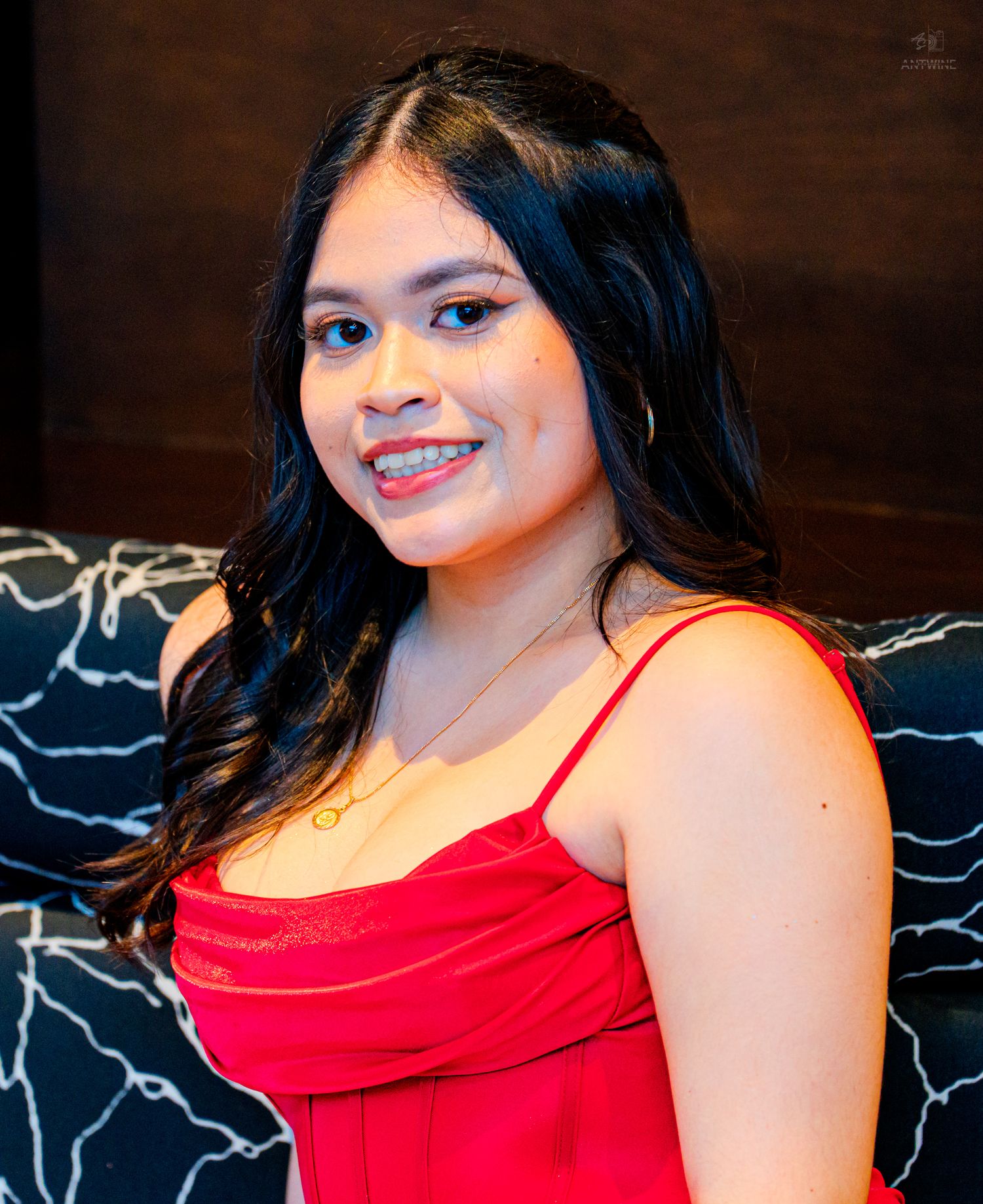 Smiling young woman with braces wearing a red dress, sitting on a dark patterned couch.