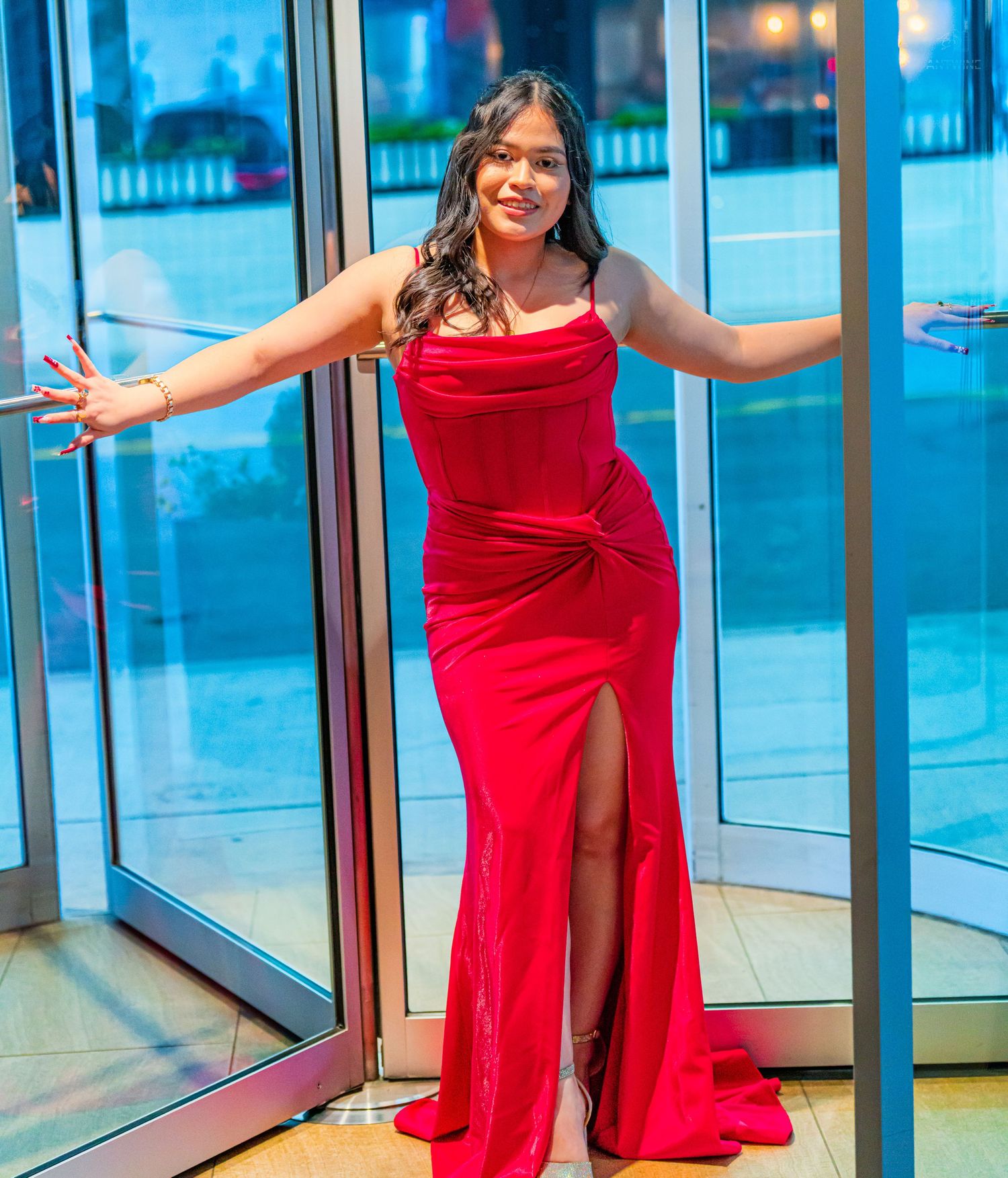 Woman in elegant red satin mermaid gown with slit poses at glass doors, smiling confidently.