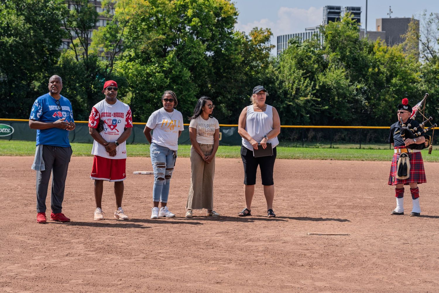 Six people and a bagpiper lined up on a softball field during a pre-game ceremony on a sunny day.