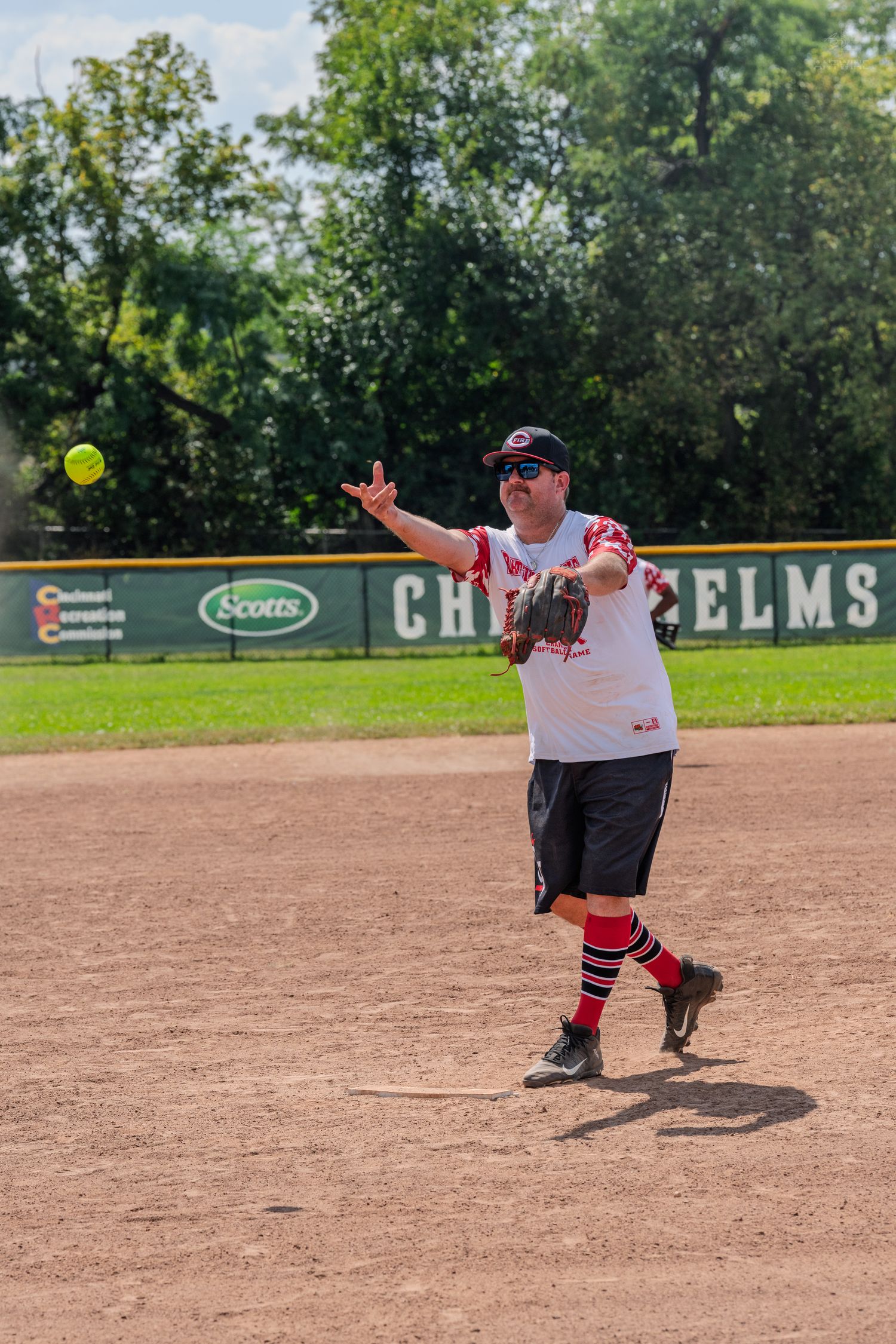 A softball player in a white and red uniform throws the ball during a sunny outdoor game at Chris Helms Field.