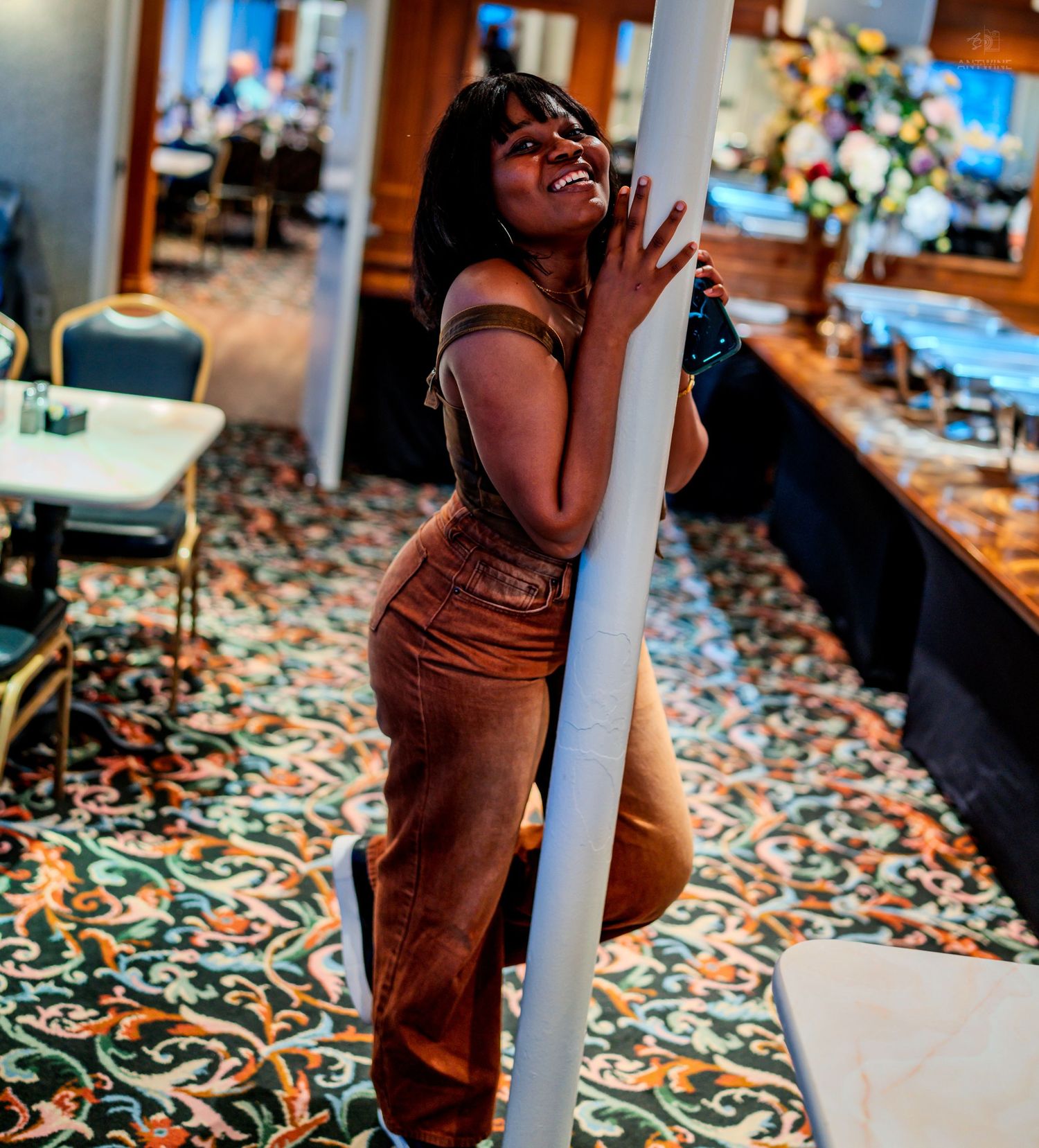 A smiling woman in brown overalls leans against a white pillar in a decorated banquet hall.
