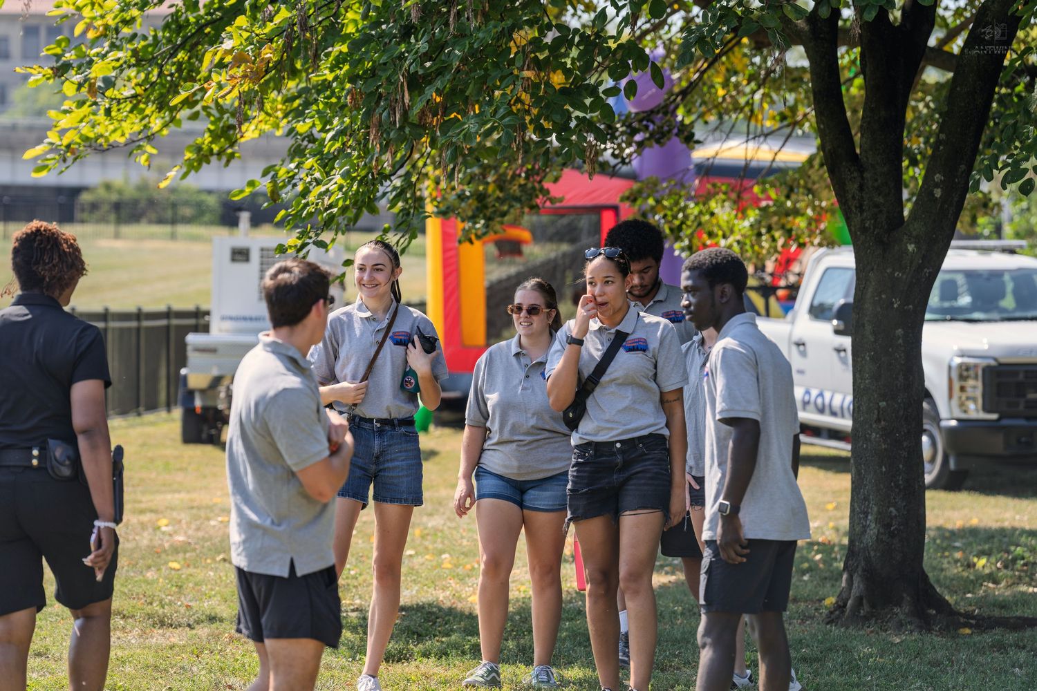 Group of young adults in matching gray shirts gathering outdoors under a tree at a sunny community event.