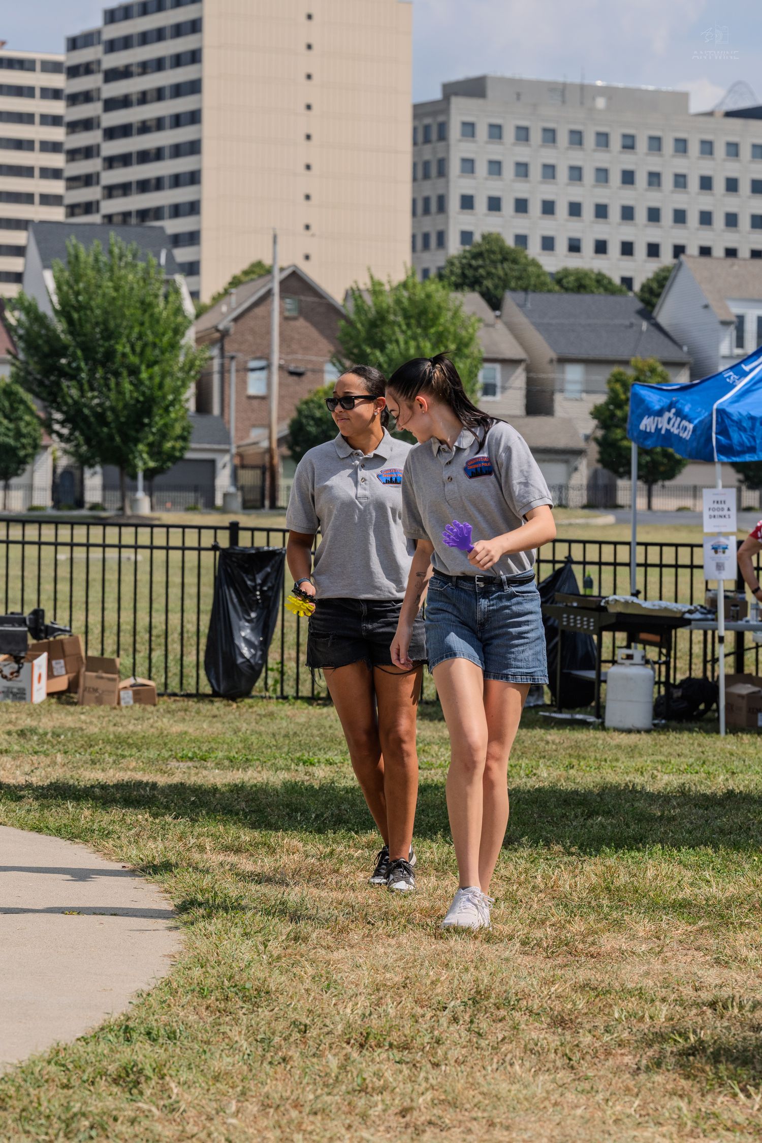 Two women in matching gray polo shirts walk together in a sunny urban park with city buildings in the background.