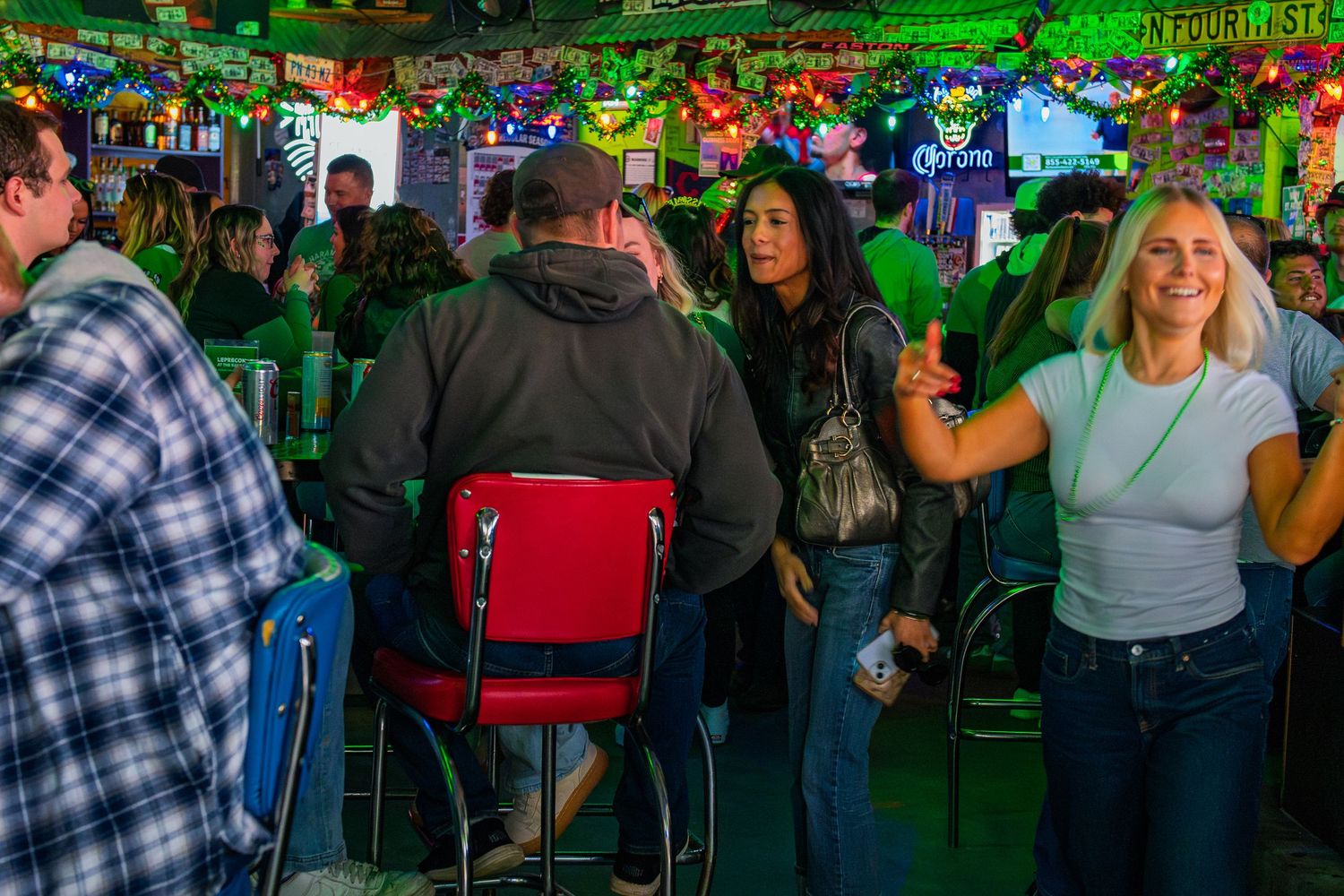 Lively bar scene with people socializing, red bar stools, and green neon lights during a festive St. Patrick's Day celebration.