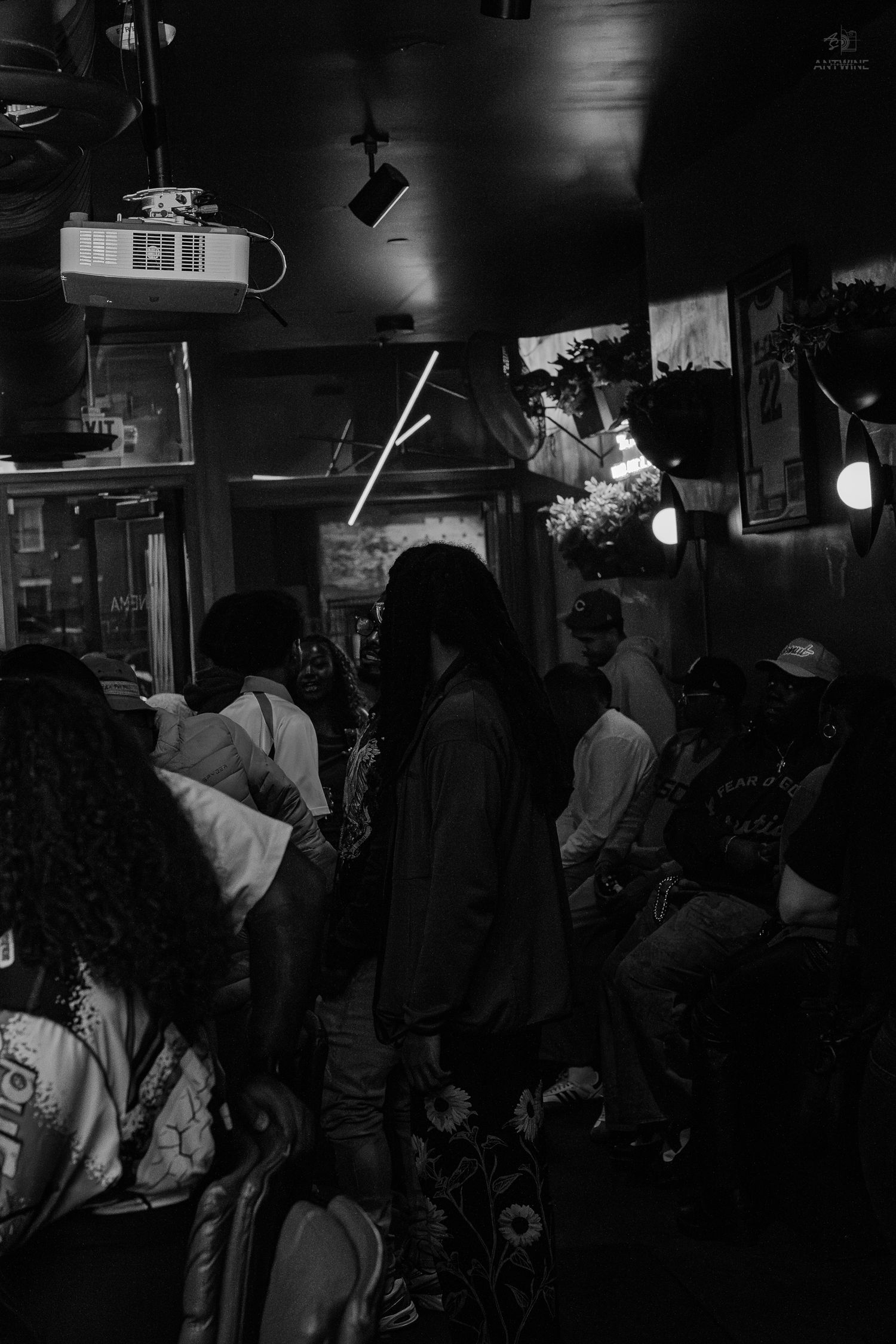 Black and white photo of a crowded indoor venue with people gathered under dim lights and a projector.